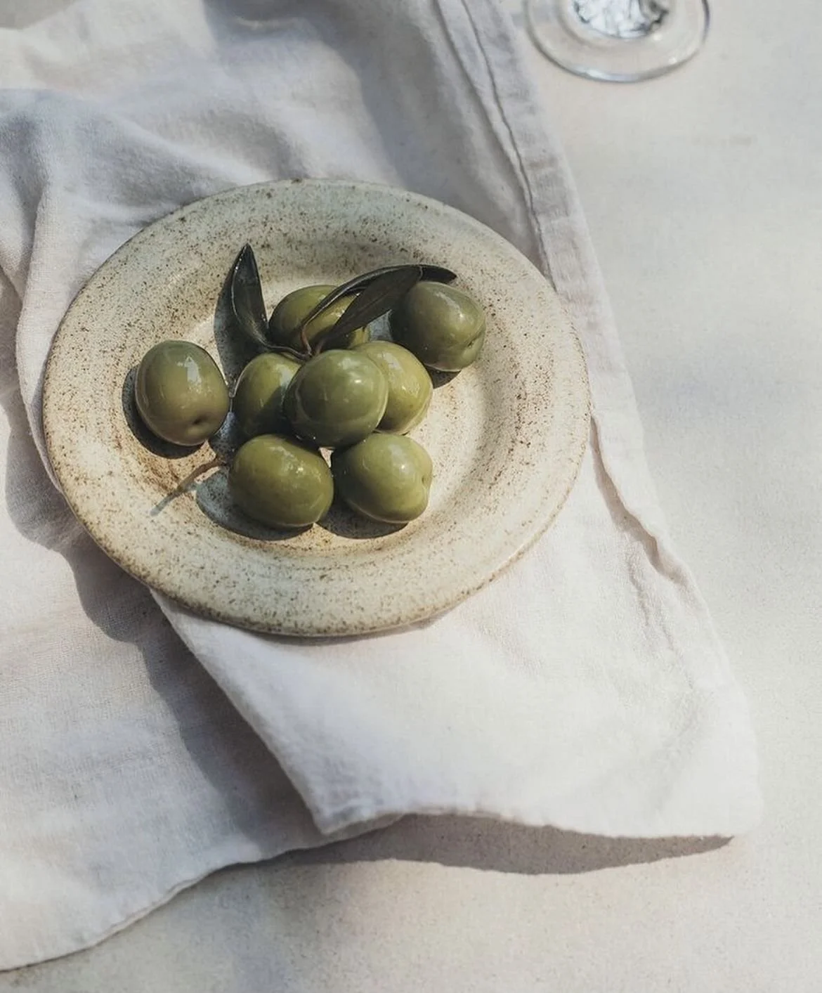 A small round plate with green olives and a few dark leaves, placed on a white cloth surface.
