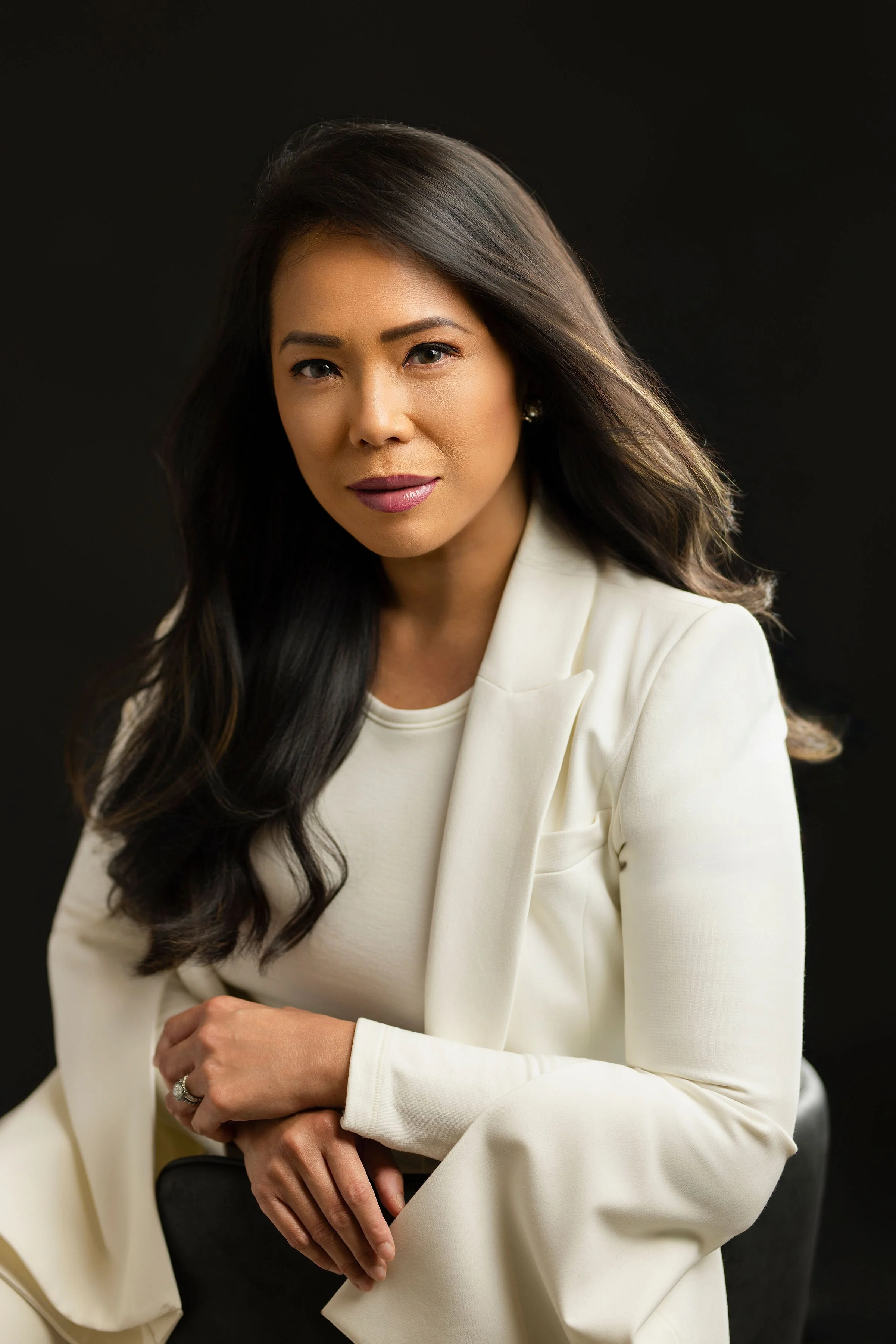 A woman with long dark hair wearing a white blazer and sitting against a black background.