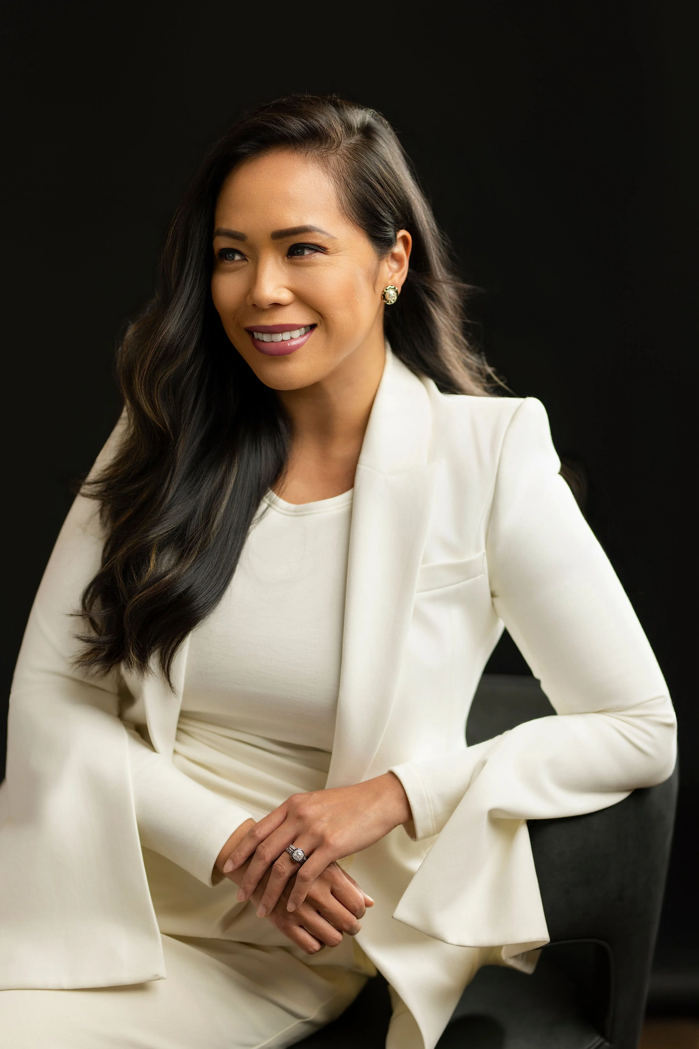 A woman with long dark hair in a white suit sitting on a chair against a black background.