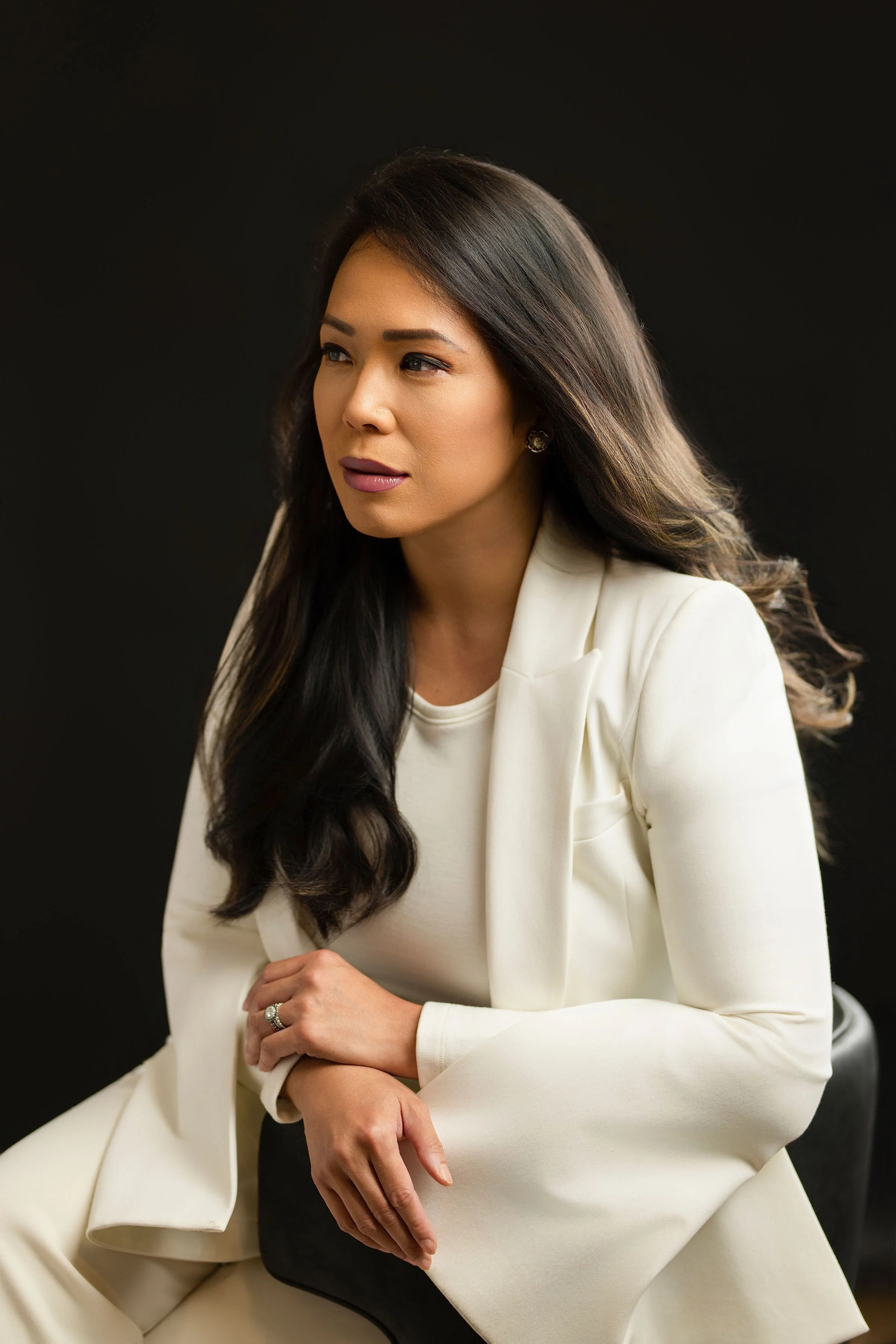 A woman with long dark hair, wearing a white blazer and a white top, sitting against a black background.