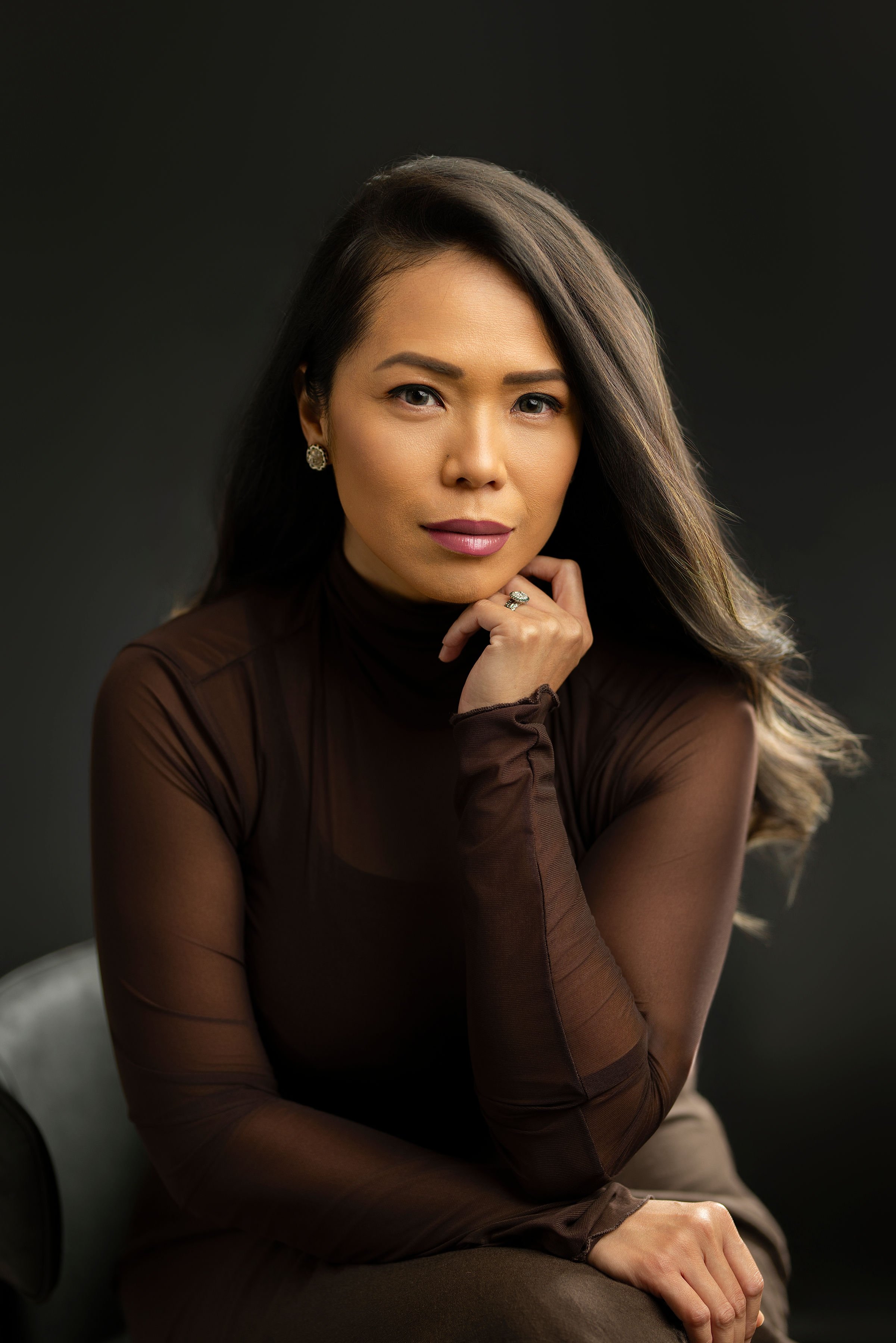 Portrait of a woman with long brown hair wearing a brown sheer top, sitting and gazing at the camera with a confident expression.