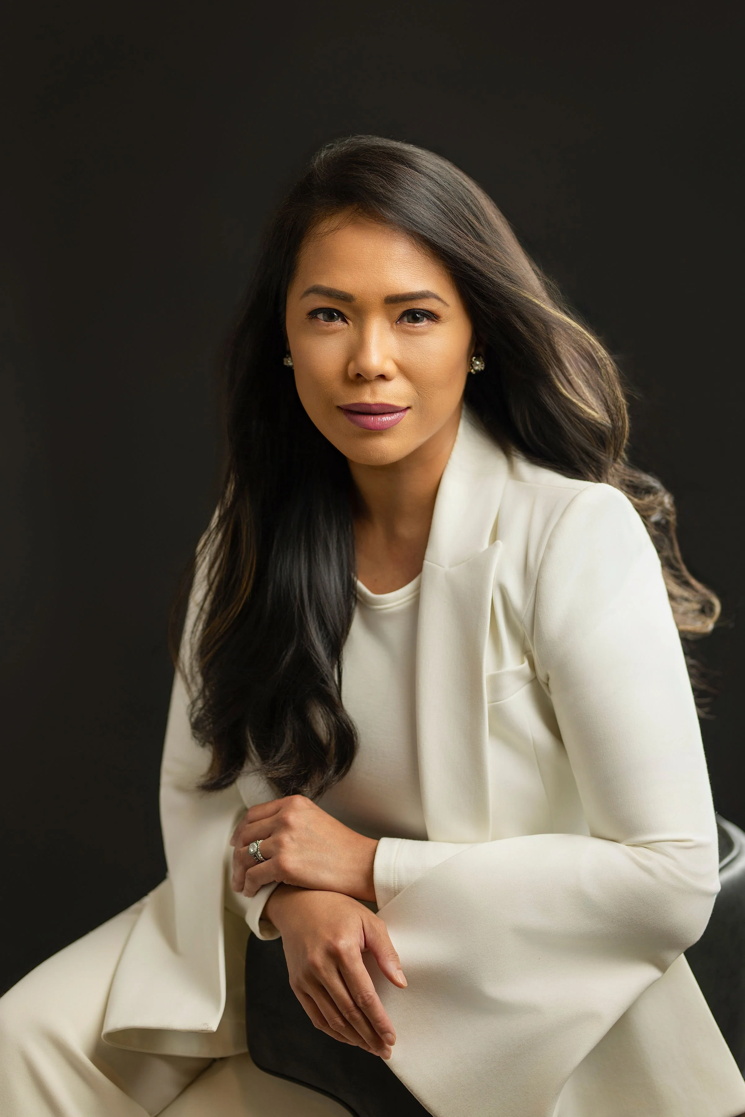 A woman with long dark hair wearing a cream-colored blazer and matching pants, sitting on a black chair against a dark background.