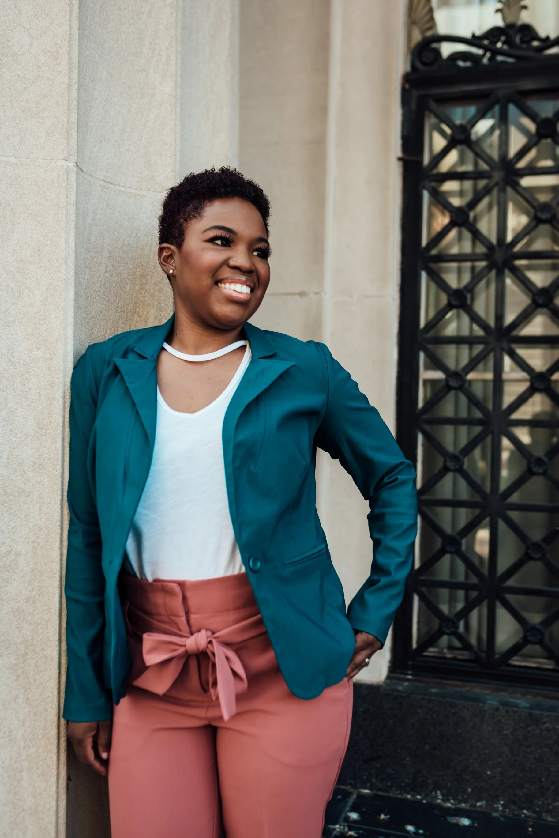 A woman with short curly hair, wearing a teal blazer, white shirt, and pink pants with a bow, smiling and leaning against a beige wall near a black decorative gate.