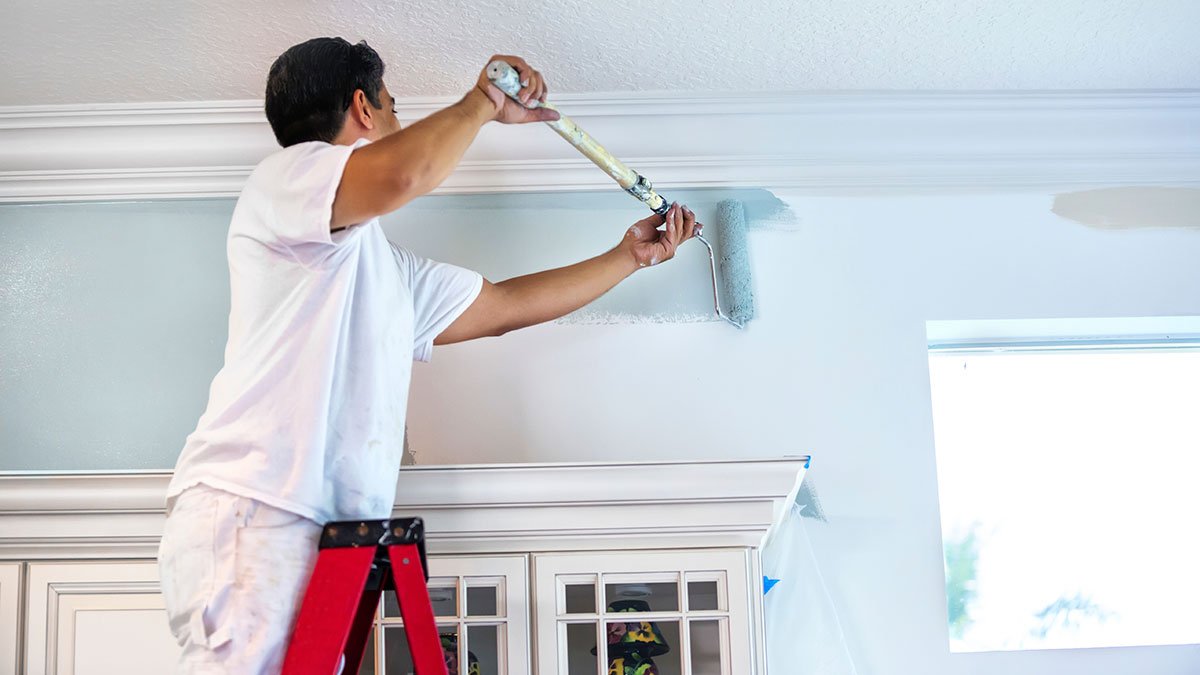 A person painting a ceiling with a roller brush, standing on a ladder in a room with a cabinet and a window.