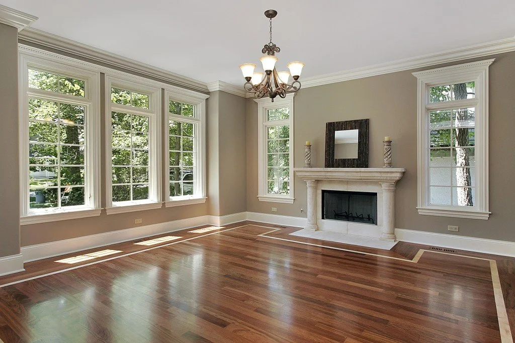 Empty living room with large windows, hardwood floor, white fireplace mantle, mirror and candleholders on the mantle, chandelier, and view of trees outside.