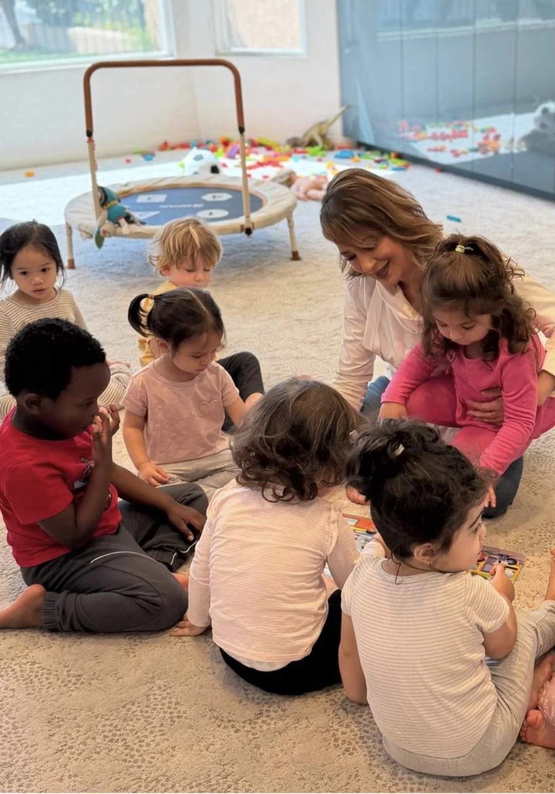 A group of young children sitting on a carpeted floor around a woman, engaging with small pictures or cards, in a bright room with toys and a playpen in the background.