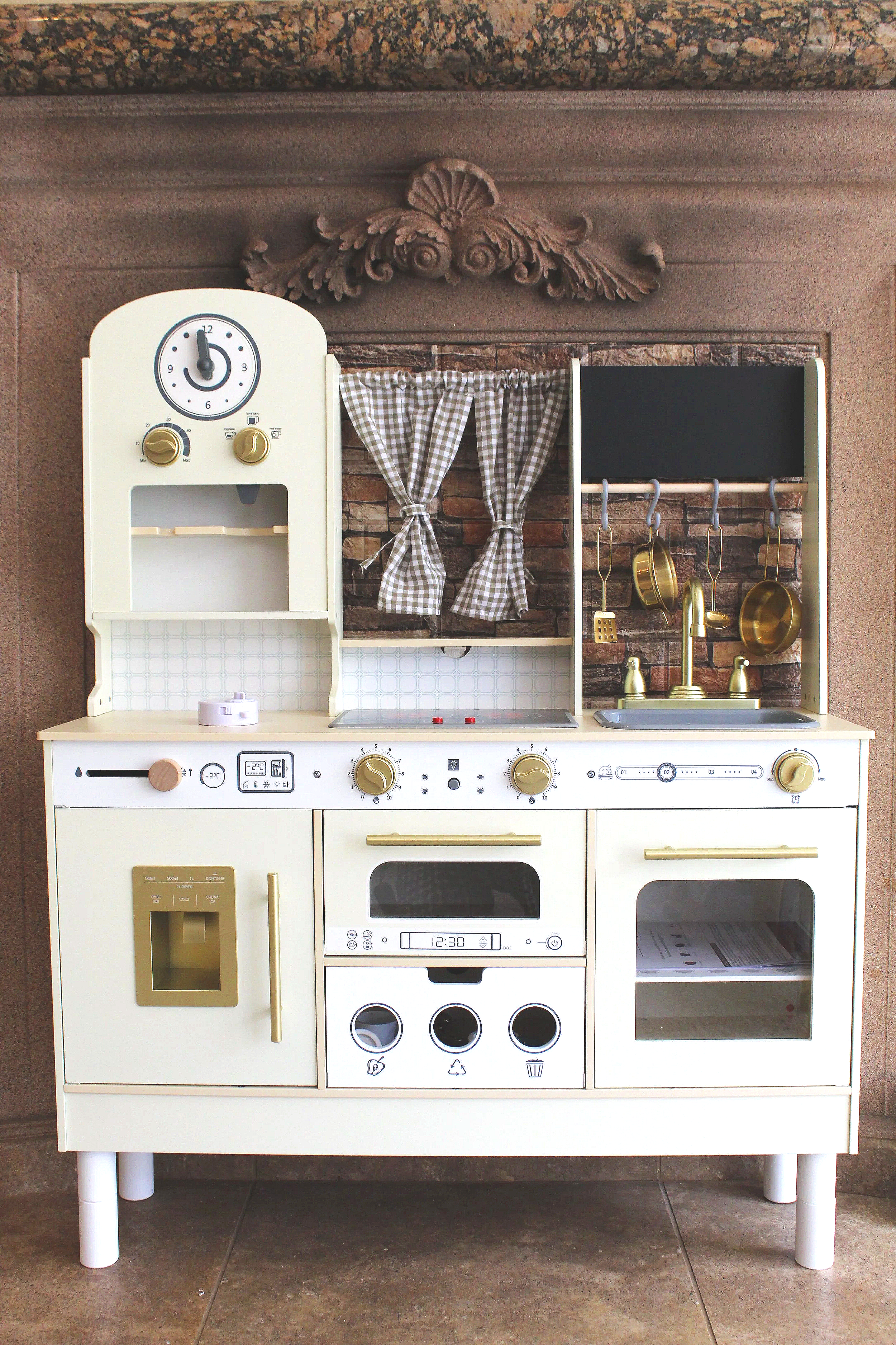 Miniature white play kitchen with gold handles, knobs, and faucet, featuring a stove, microwave, oven, and sink. Decorative checkered curtains and hanging pots are also visible.