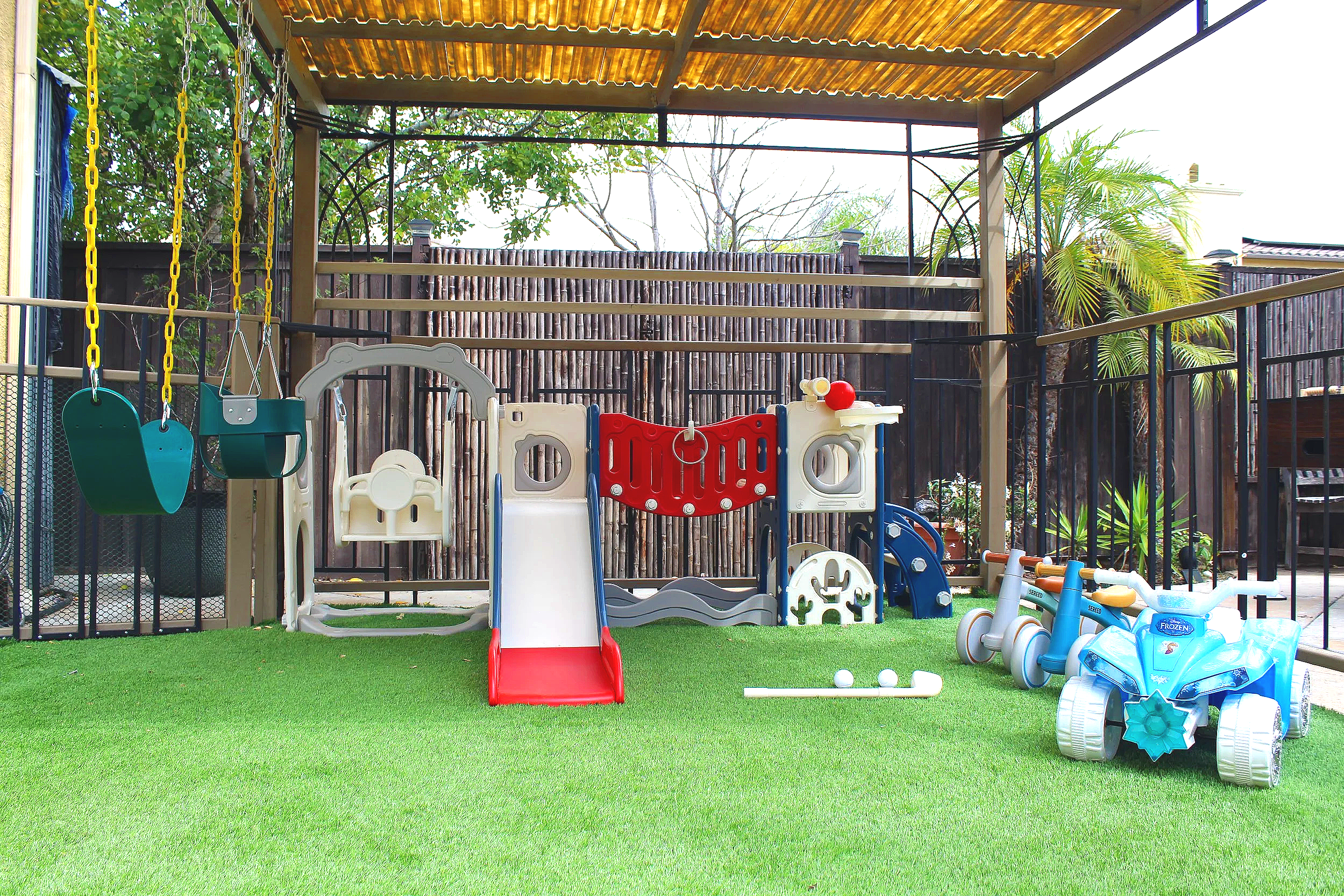 Children's outdoor play area with swings, slides, and ride-on toy cars on artificial grass, enclosed by a wooden fence and partial roof.