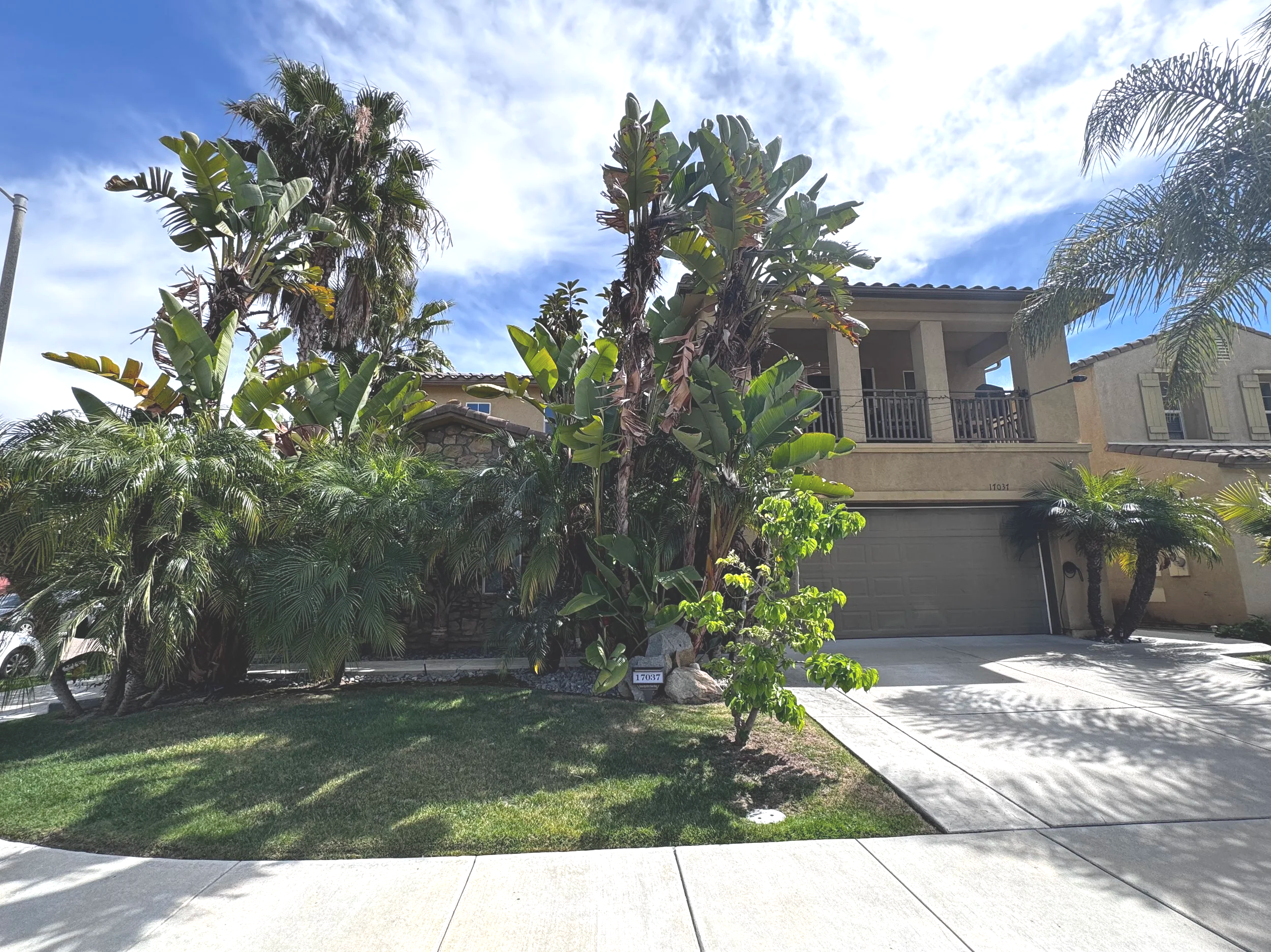 A two-story house with a beige exterior, a balcony, and a garage, surrounded by palm trees and lush greenery under a partly cloudy sky.