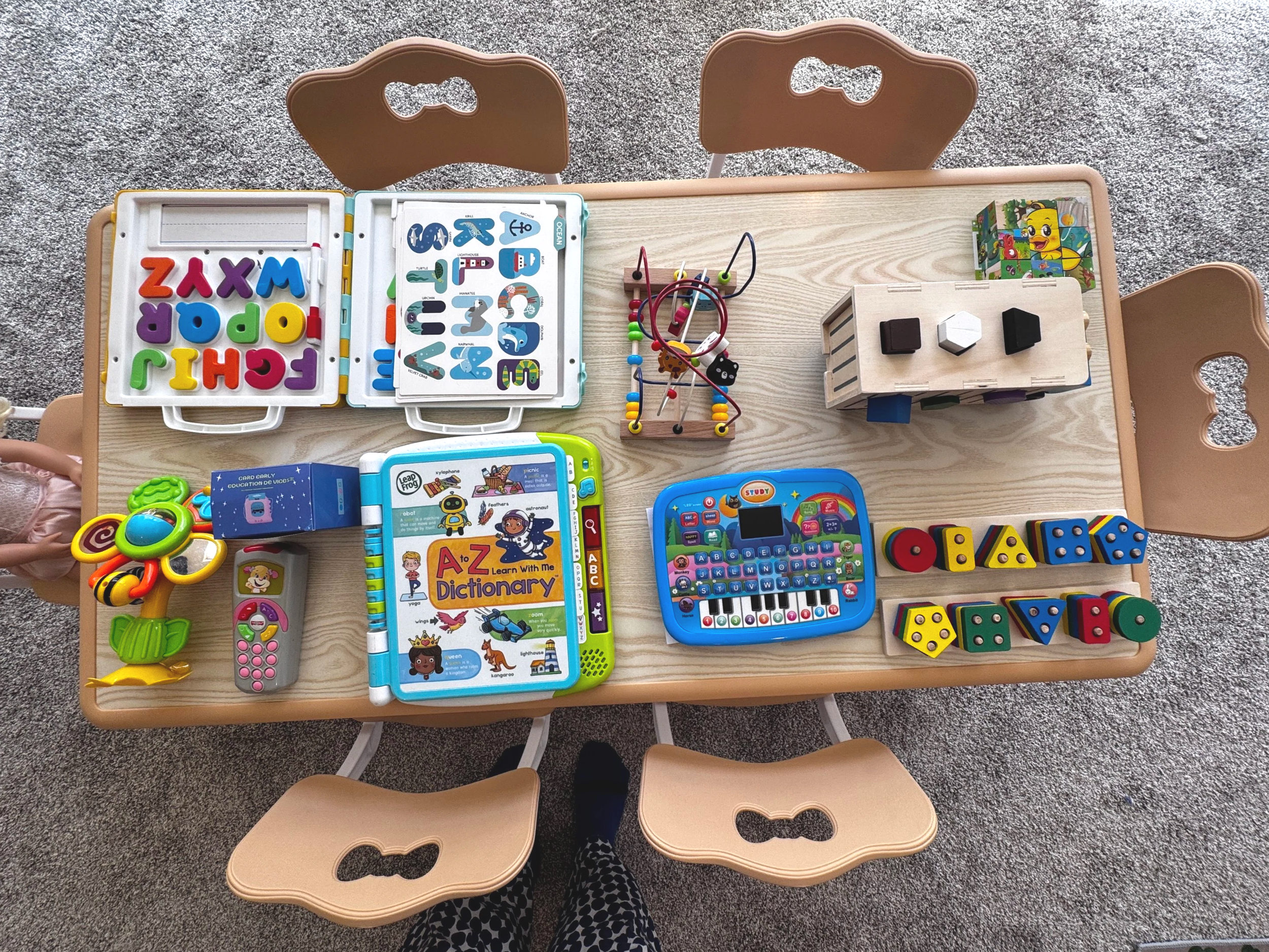 Toy table with educational and play items including alphabet and number flashcards, magnetic letters, a bead maze, wooden block puzzles, a shape and color matching toy, a toy keyboard, a flower-shaped rattle, a remote control, and a box of cards, on 