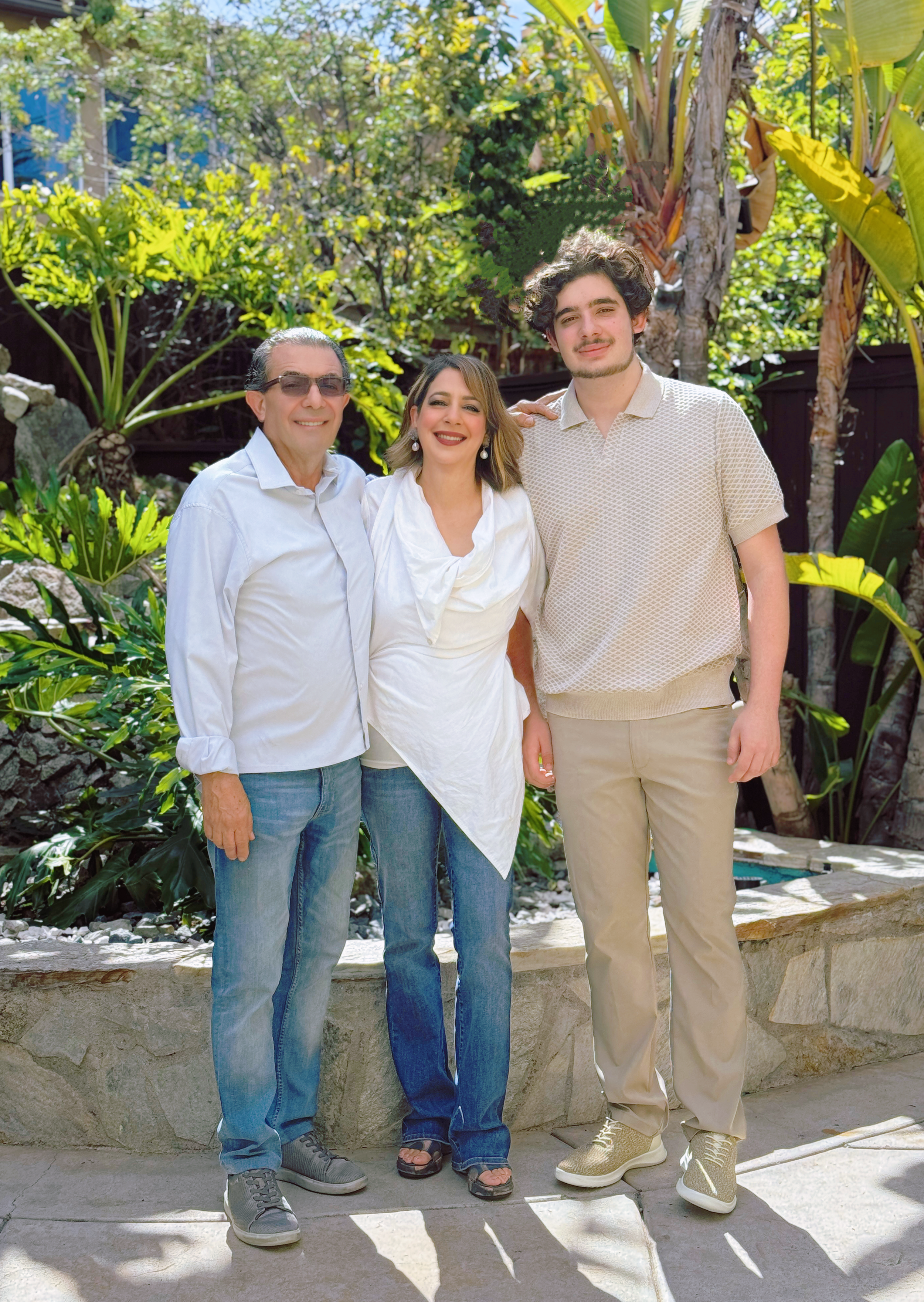 A family of three standing together outdoors in a lush garden, smiling at the camera. The family includes a man with gray hair wearing sunglasses, a woman with shoulder-length brown hair, and a young man with curly dark hair. The group is dressed casually, with the man and woman in white tops and the young man in a light-colored polo shirt. The background features dense greenery and tall tropical plants.
