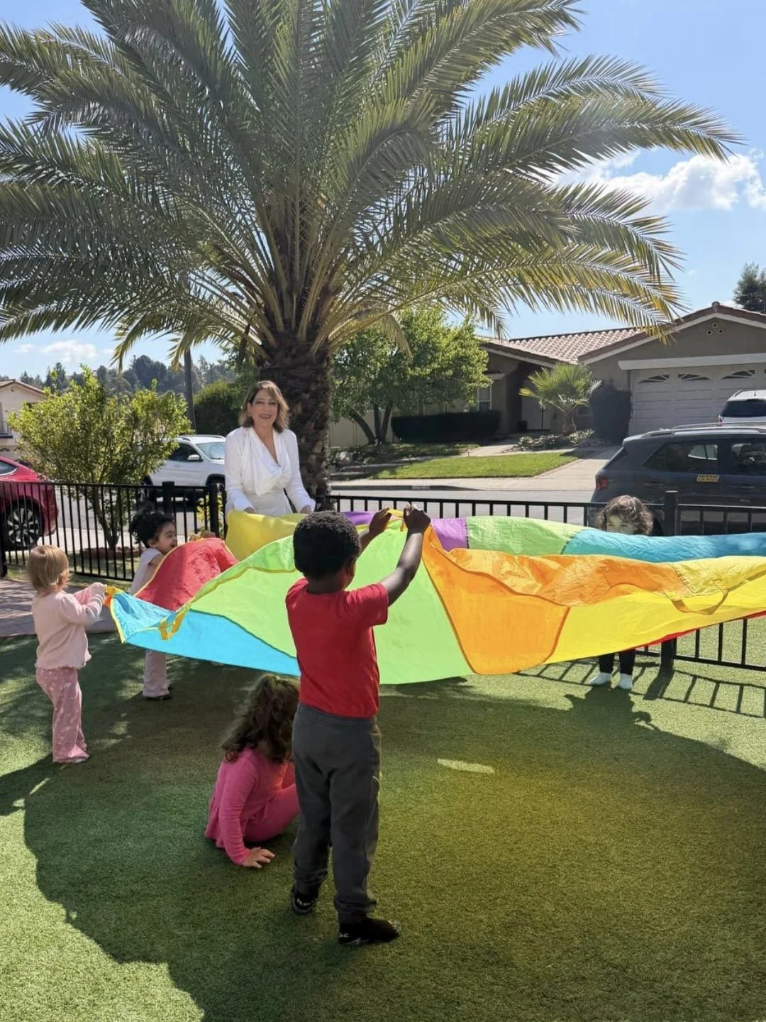 Children and an adult holding a colorful parachute outside on a sunny day, with a large palm tree, residential houses, and a parked car in the background.
