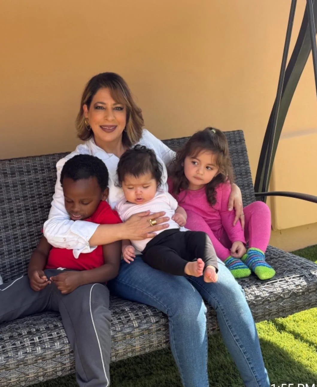 A woman sitting on a woven outdoor bench with four young children, holding the youngest on her lap and another close to her, on a sunny day in front of a yellow wall.
