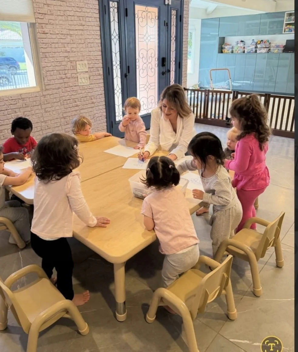 A group of young children gathered around a table in a classroom, with a teacher engaging with them. The children are drawing or coloring on sheets of paper, and the classroom has bright lighting and colorful storage units in the background.
