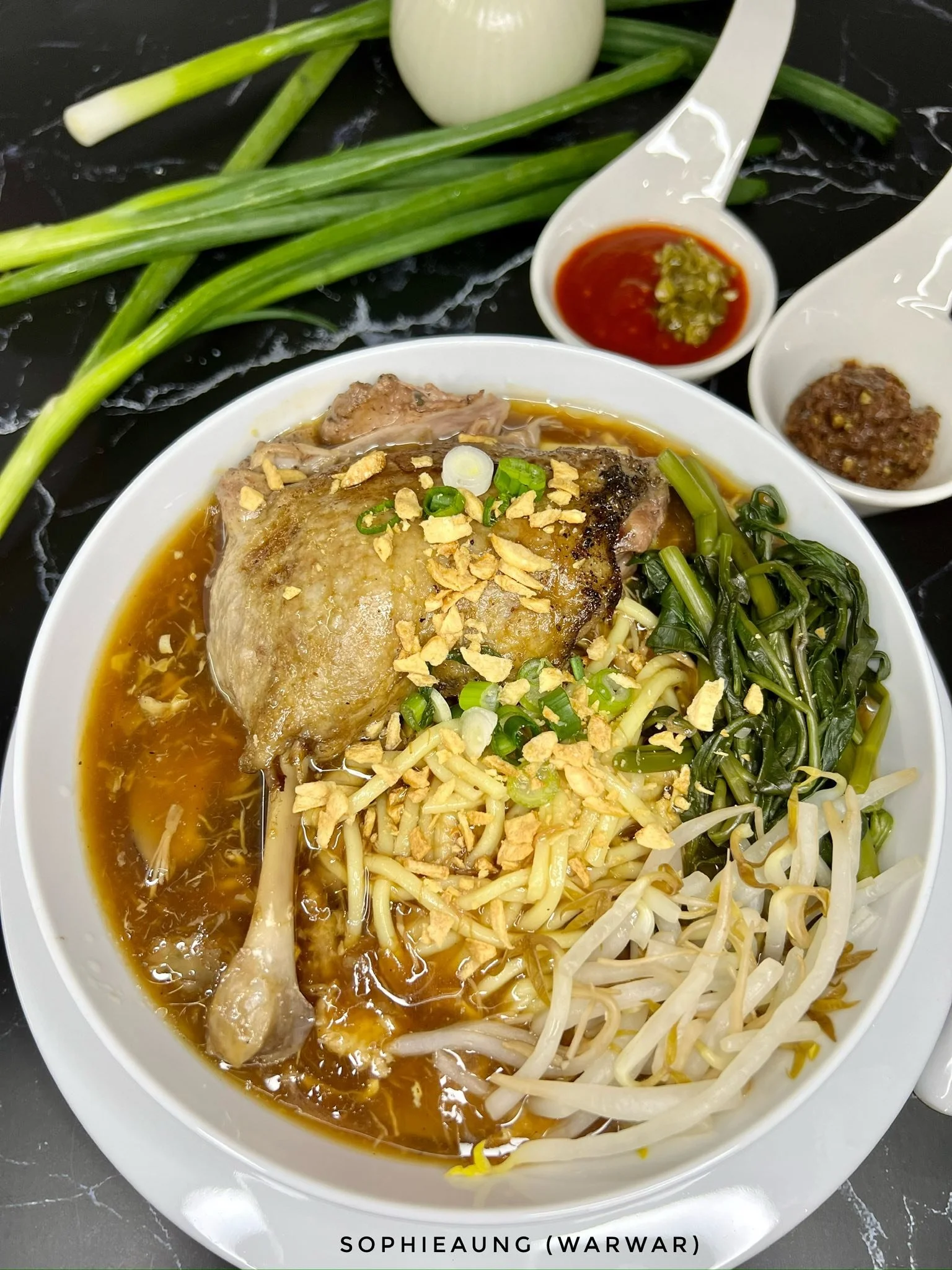 A bowl of ramen with chicken, green onions, bean sprouts, and vegetables, topped with garlic crisps, served with side sauces and fresh green onions on a black marble surface.