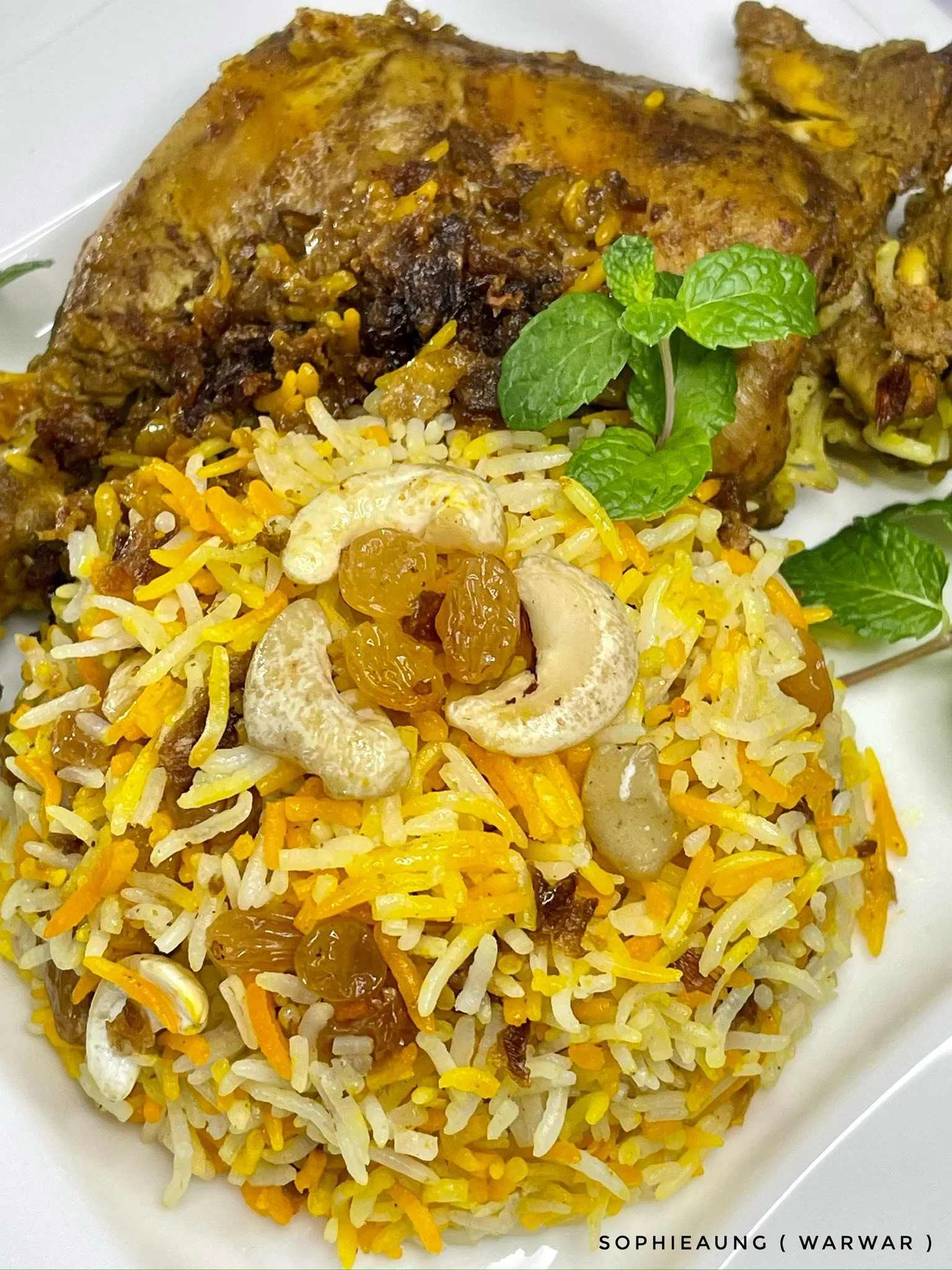 A plate of Indian-style rice with cashews, golden raisins, and spices, garnished with a sprig of mint, served with a portion of cumin-spiced chicken.