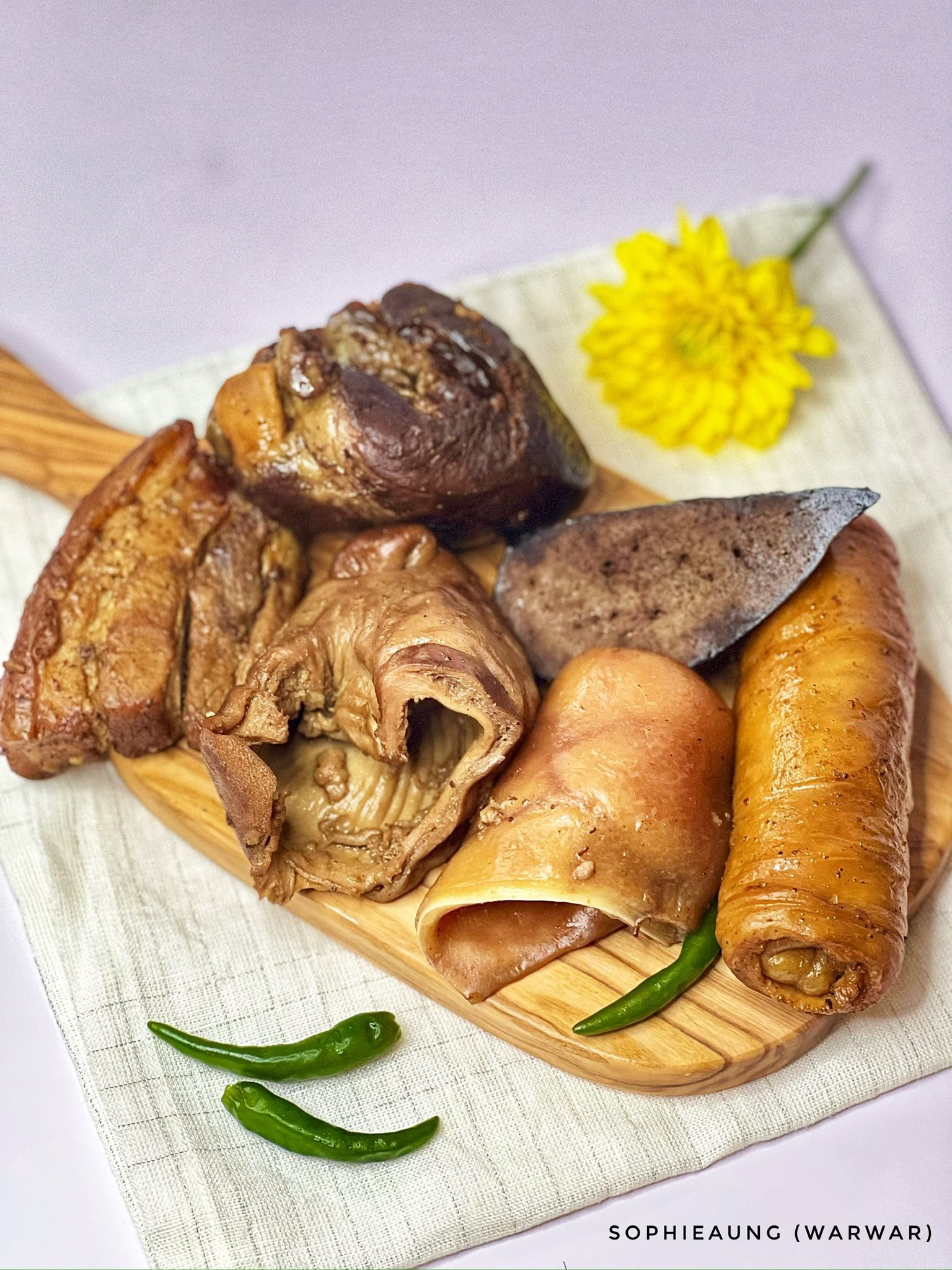 Assorted cooked pig parts including ears, liver, intestine, and other cuts on a wooden platter with a yellow flower and green chili peppers.