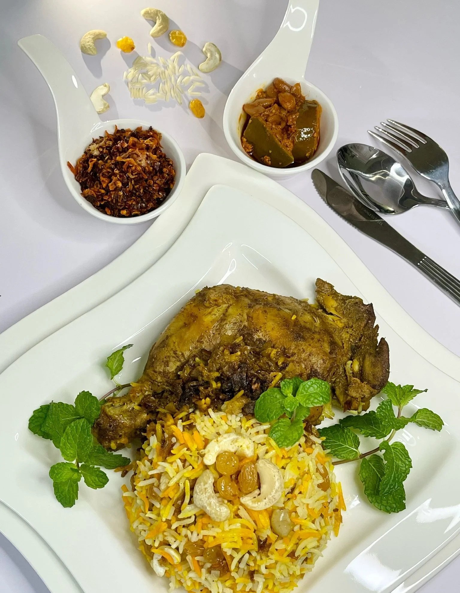 Plate with cooked rice garnished with cashews, raisins, and herbs, and a piece of cooked meat. Two small bowls of spicy vegetable dishes are at the top, with the bowl on the left containing a dark, chili-based mixture, and the bowl on the right containing a yellow-green stew. Silverware is arranged to the right.