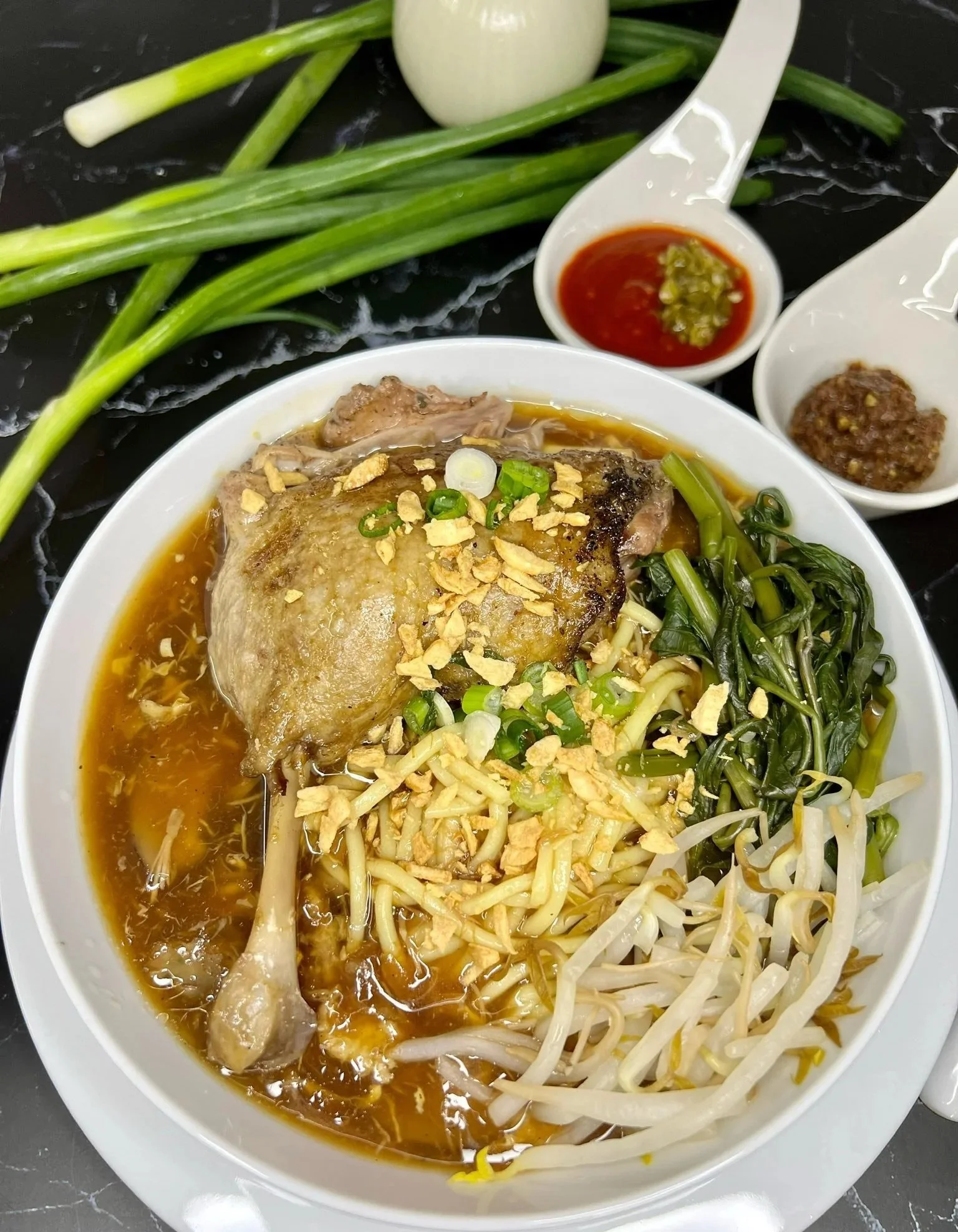 Bowl of Vietnamese pho with chicken, bean sprouts, green onions, and herbs, served with side condiments including chili sauce, hoisin sauce, and sliced garlic chili paste, with green onions and garlic on the side.
