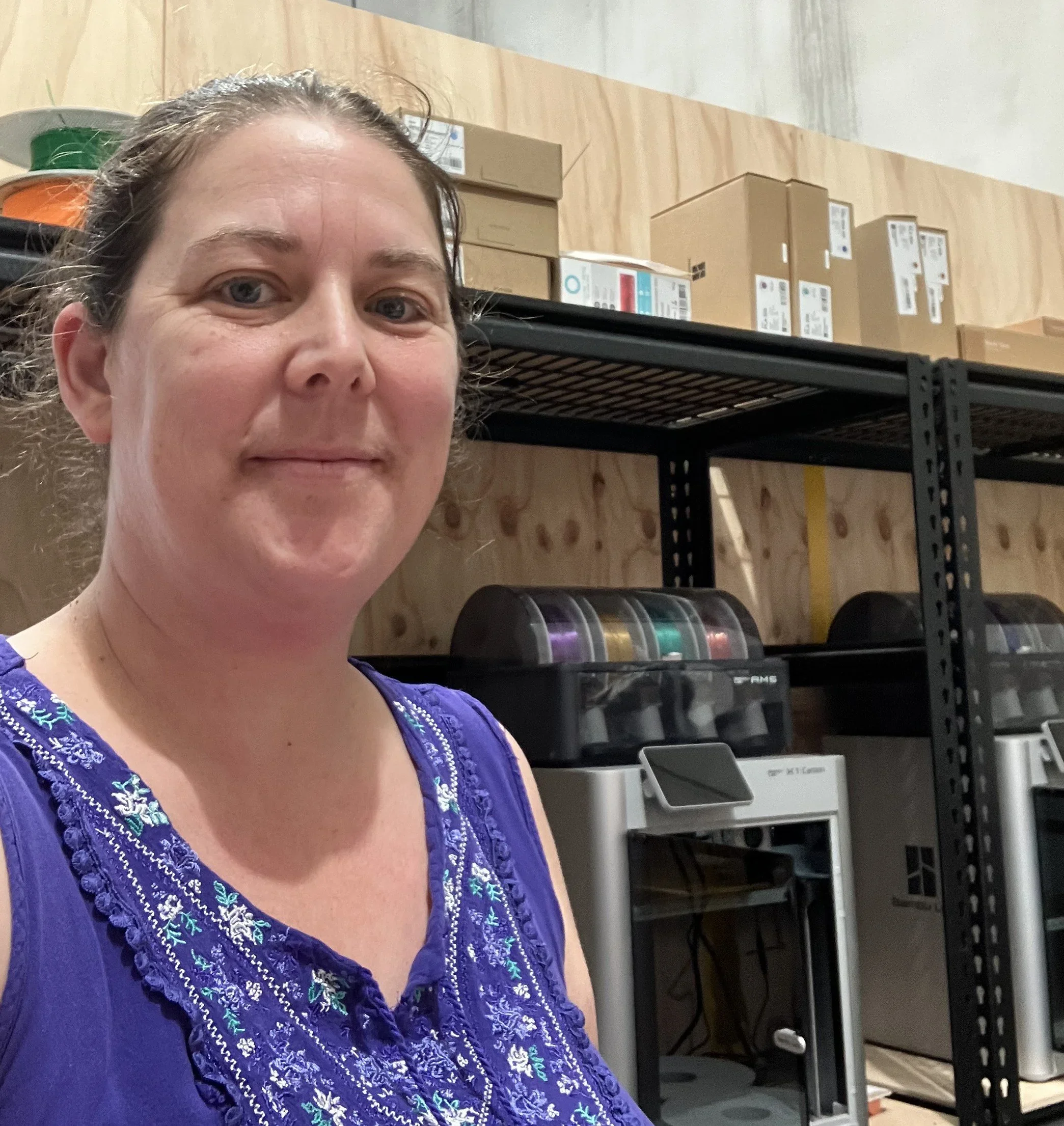 A woman with brown hair pulled back, wearing a blue embroidered top, standing in front of a storage area with cardboard boxes and printers.