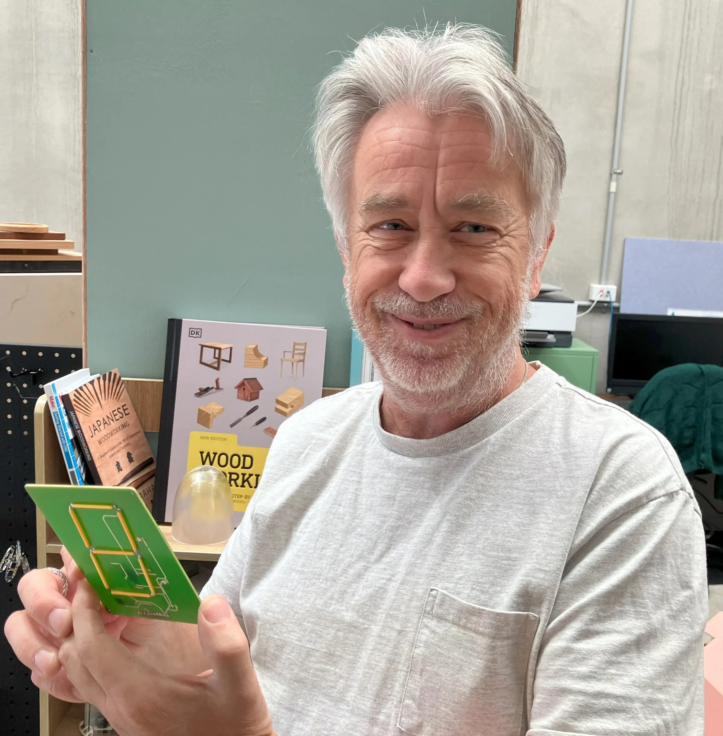 An elderly man with gray hair and a beard smiling while holding a green circuit board in a workshop or office environment with woodworking books and tools in the background.