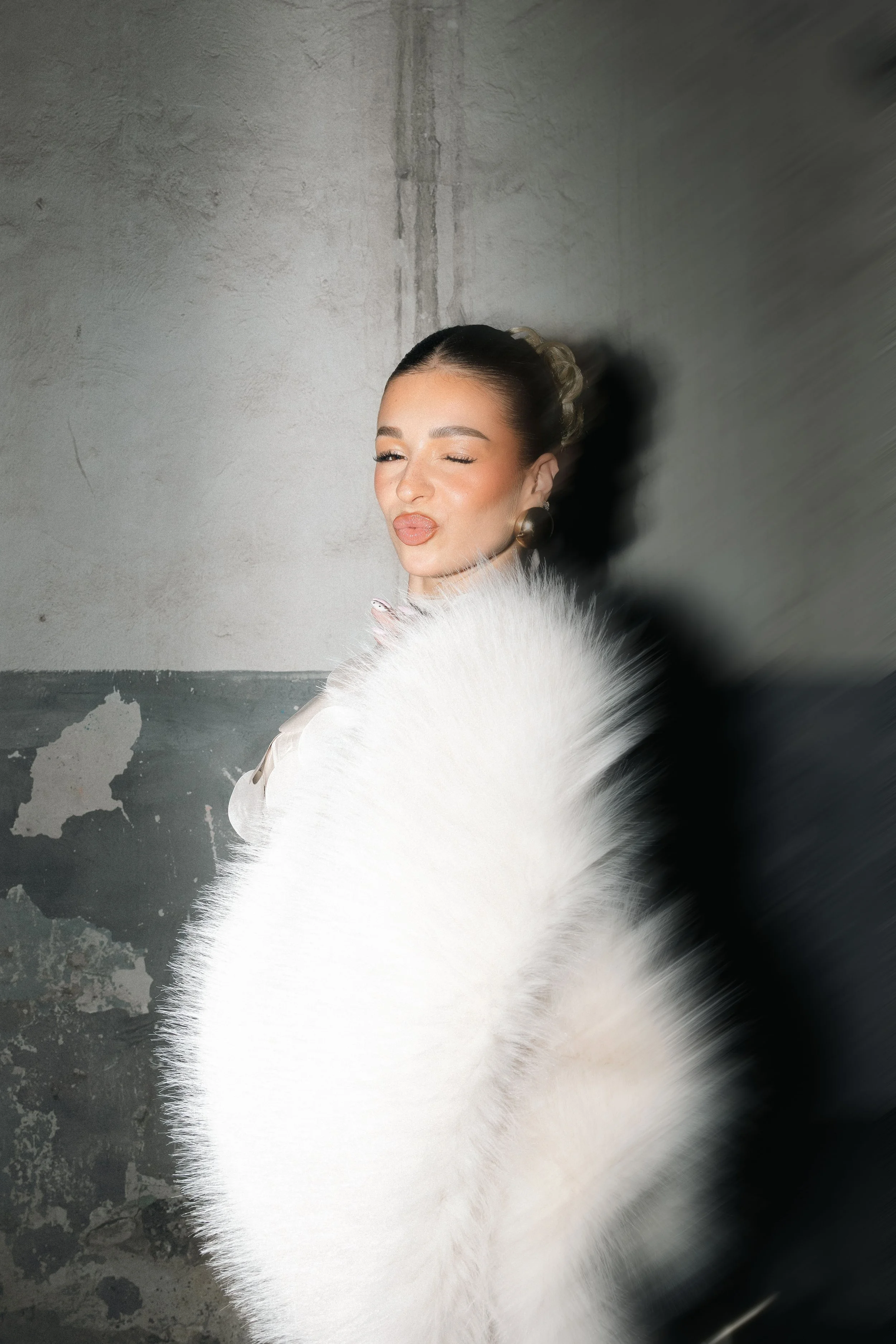 Woman posing with a playful expression, wearing large earrings and a white fur stole against a textured gray wall.