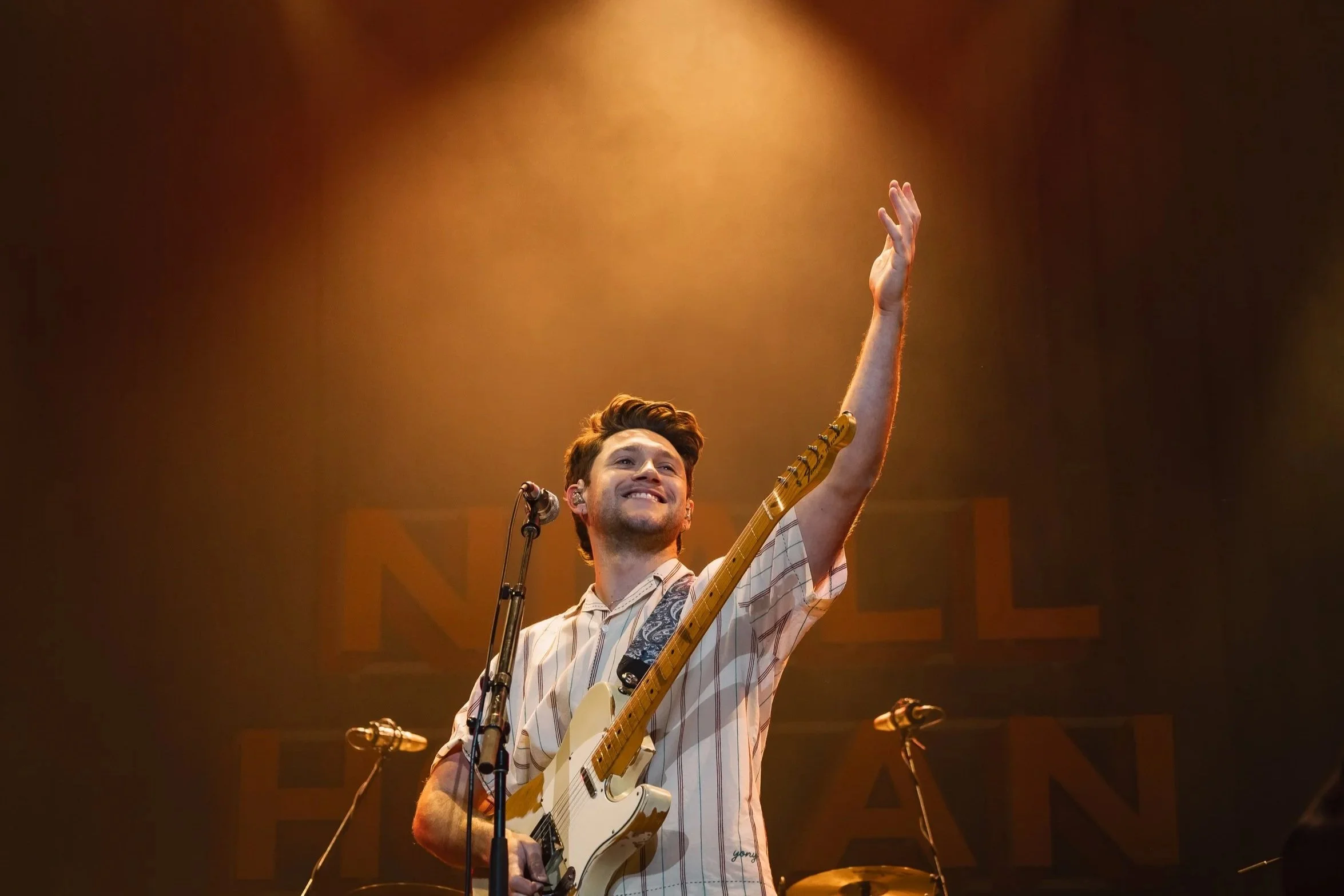 A young man with short brown hair smiling and holding a cream-colored electric guitar on stage, with his right arm raised, in front of a microphone with a black stand, under warm stage lighting.