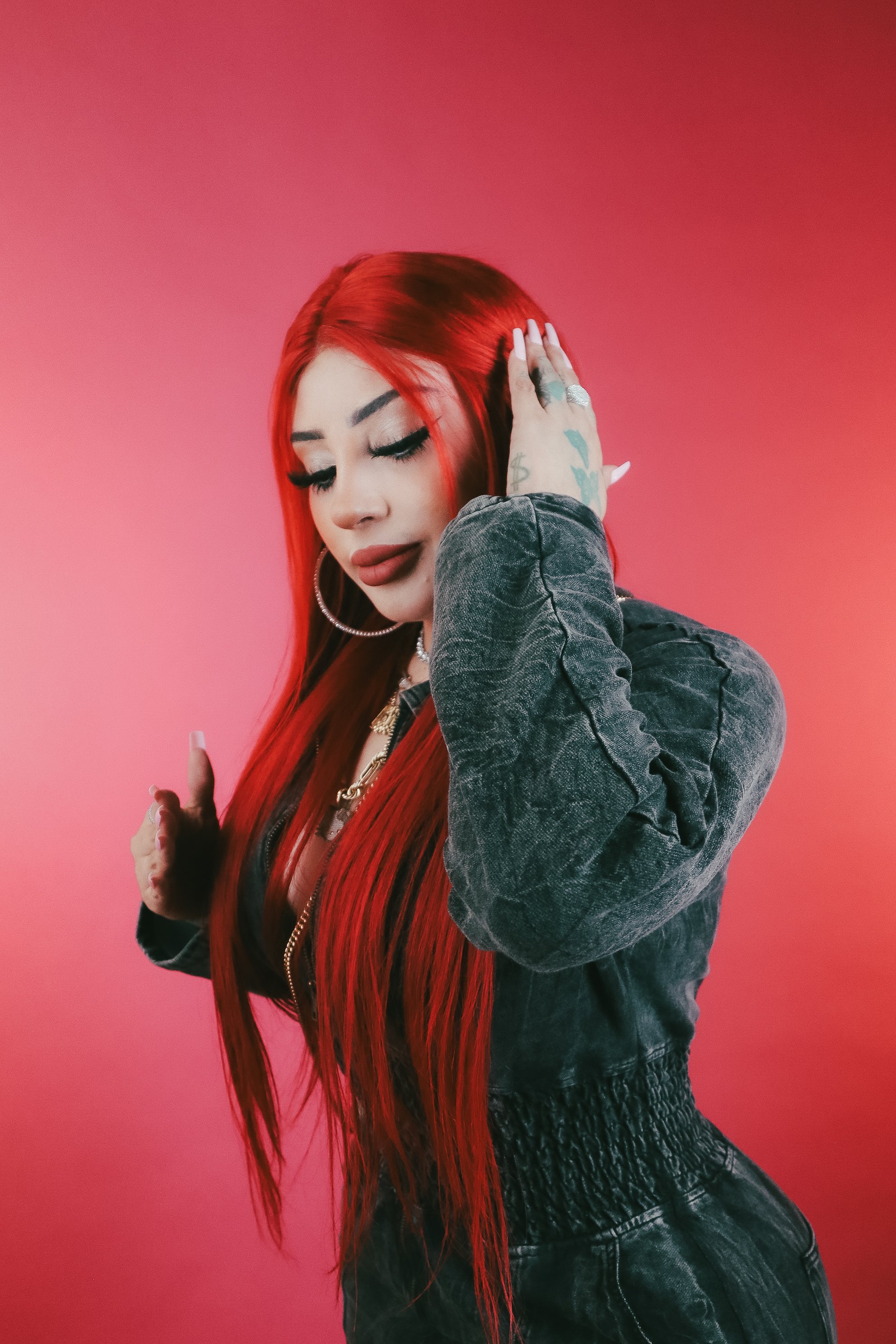 A woman with long red hair, tattoos, hoop earrings, and layered necklaces poses against a pink background, touching her ear with her left hand and wearing a black textured jacket.