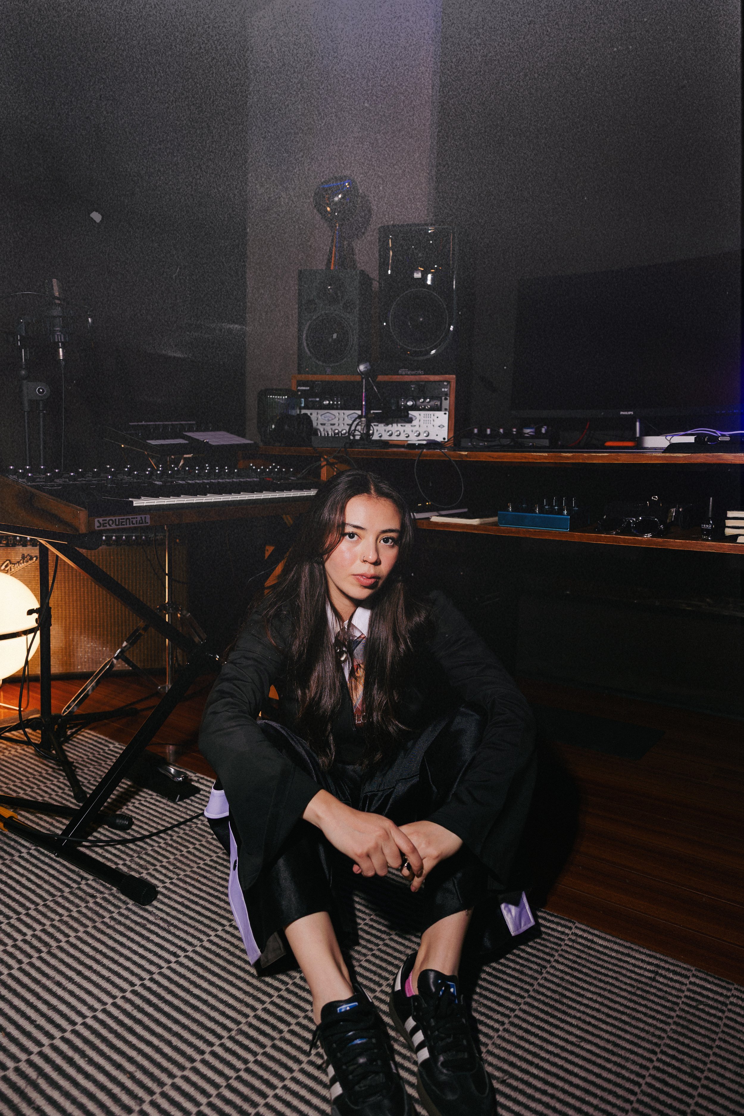 A young woman with long dark hair sitting on a patterned rug in a music studio, surrounded by music equipment, with a relaxed expression.