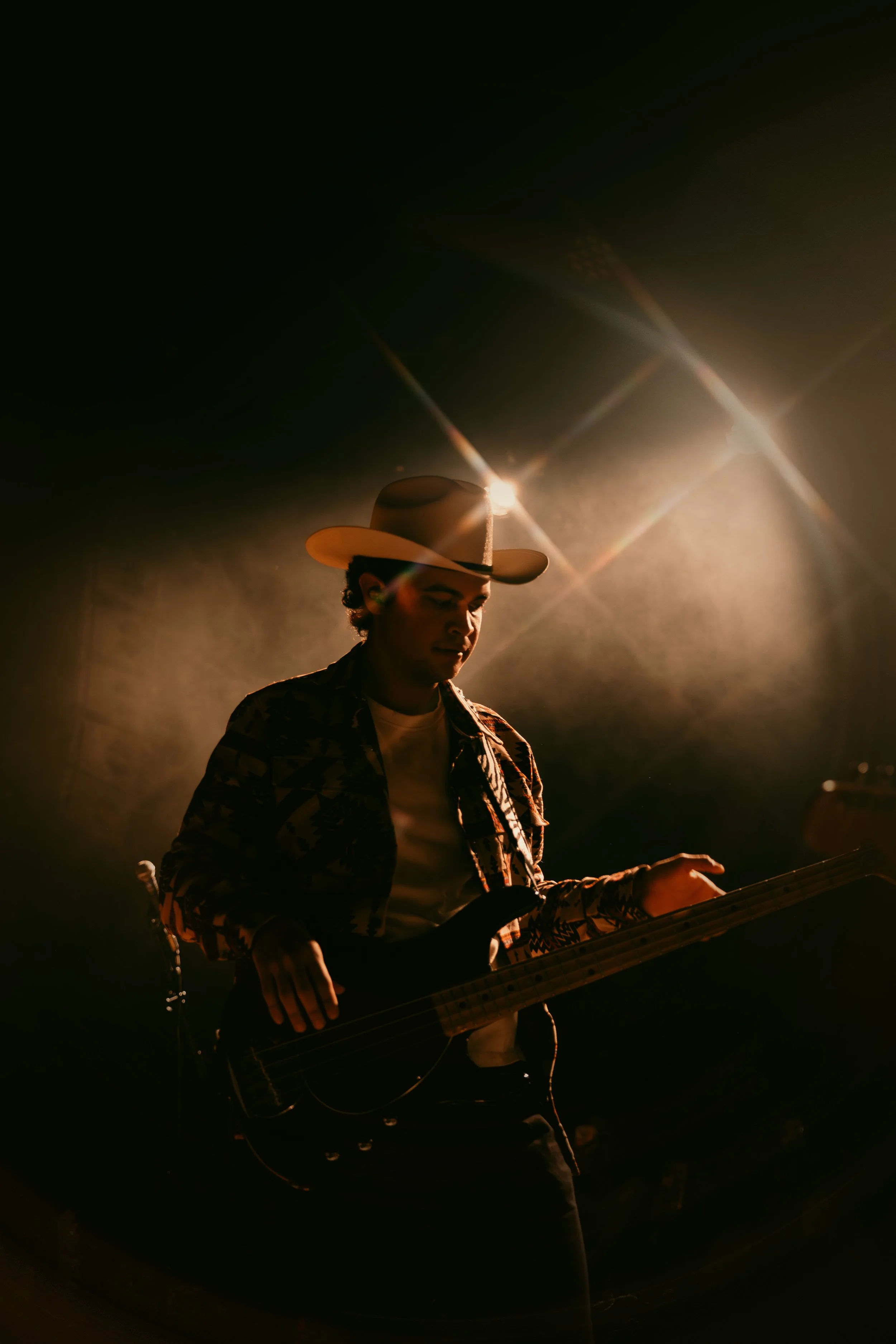 A person wearing a cowboy hat and patterned jacket playing an electric bass guitar on stage with dramatic lighting.