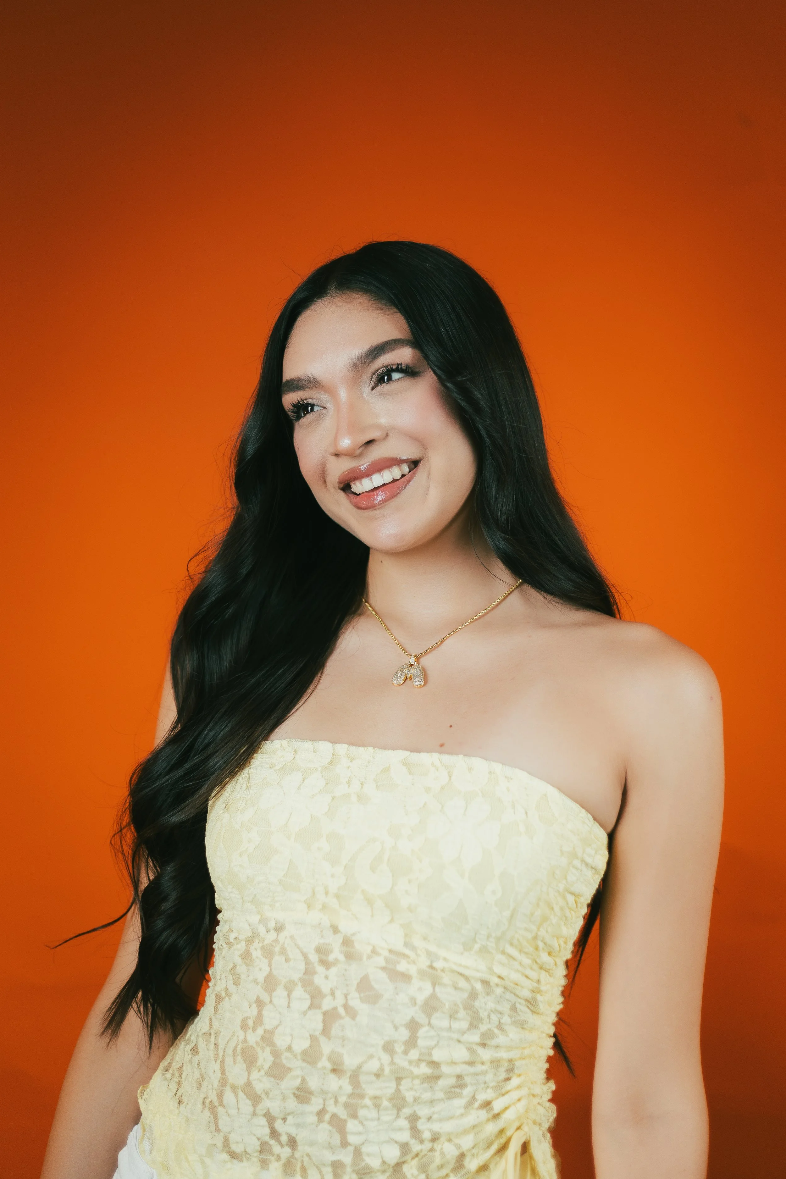 A young woman with long black hair smiling, wearing a yellow strapless lace dress and a gold necklace with a heart-shaped pendant, standing against an orange background.