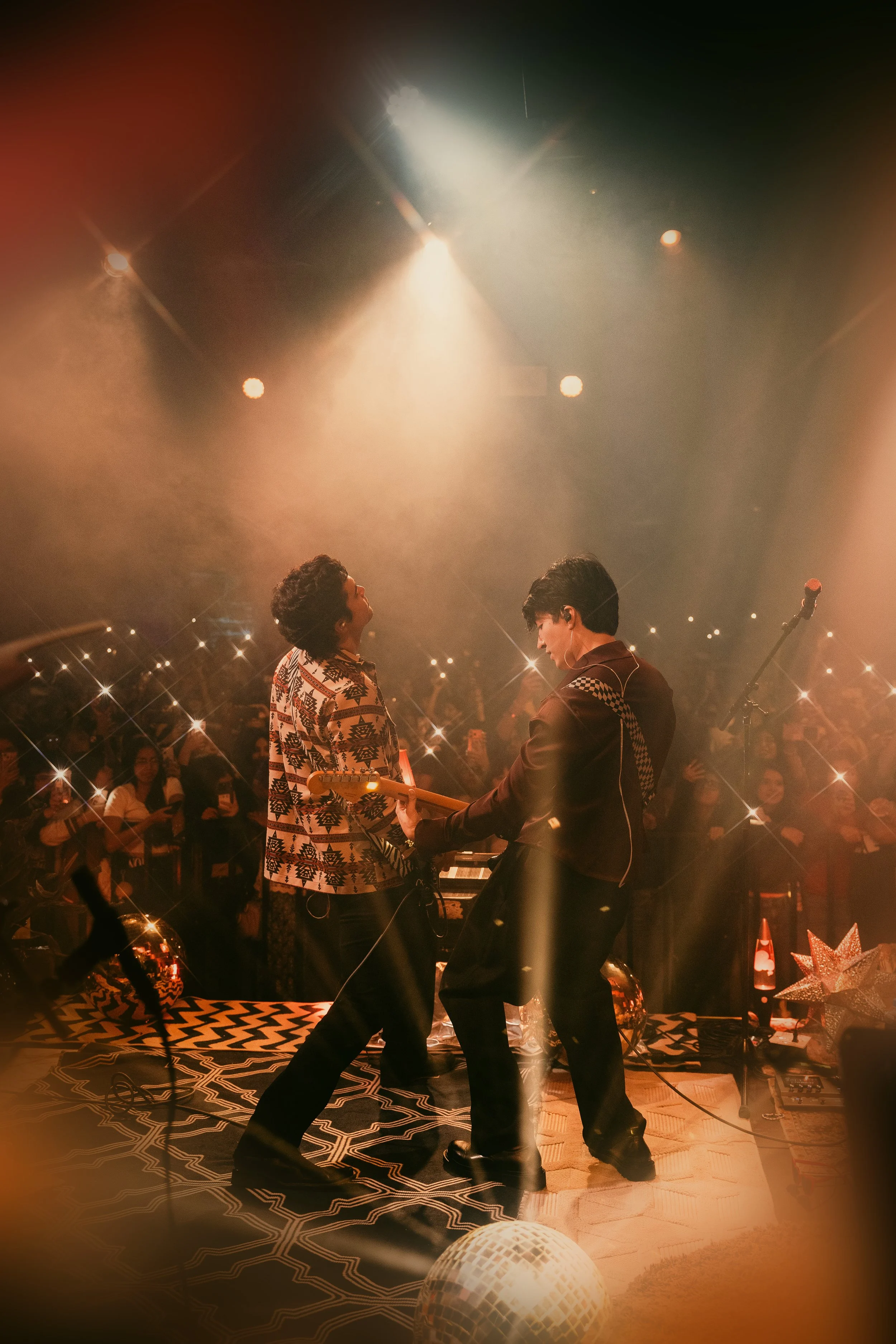 Two musicians perform on stage during a lively concert, surrounded by an enthusiastic crowd with many holding up phones and lights, under warm stage lighting.