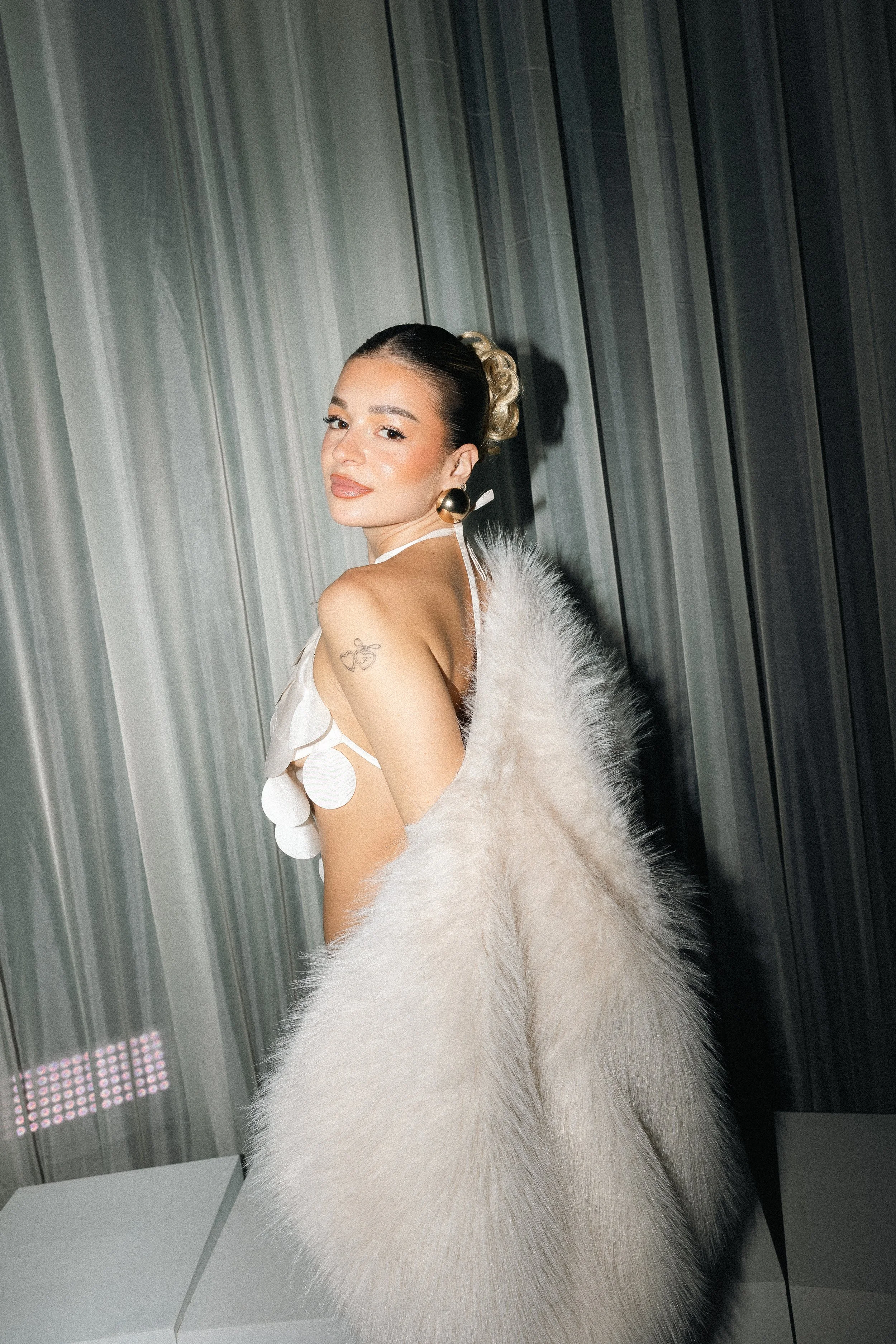 A woman dressed in a glamorous, elegant outfit with large gold earrings, a white fur coat draped over her shoulders, and a unique top with circular patterns, standing against a metallic wall.