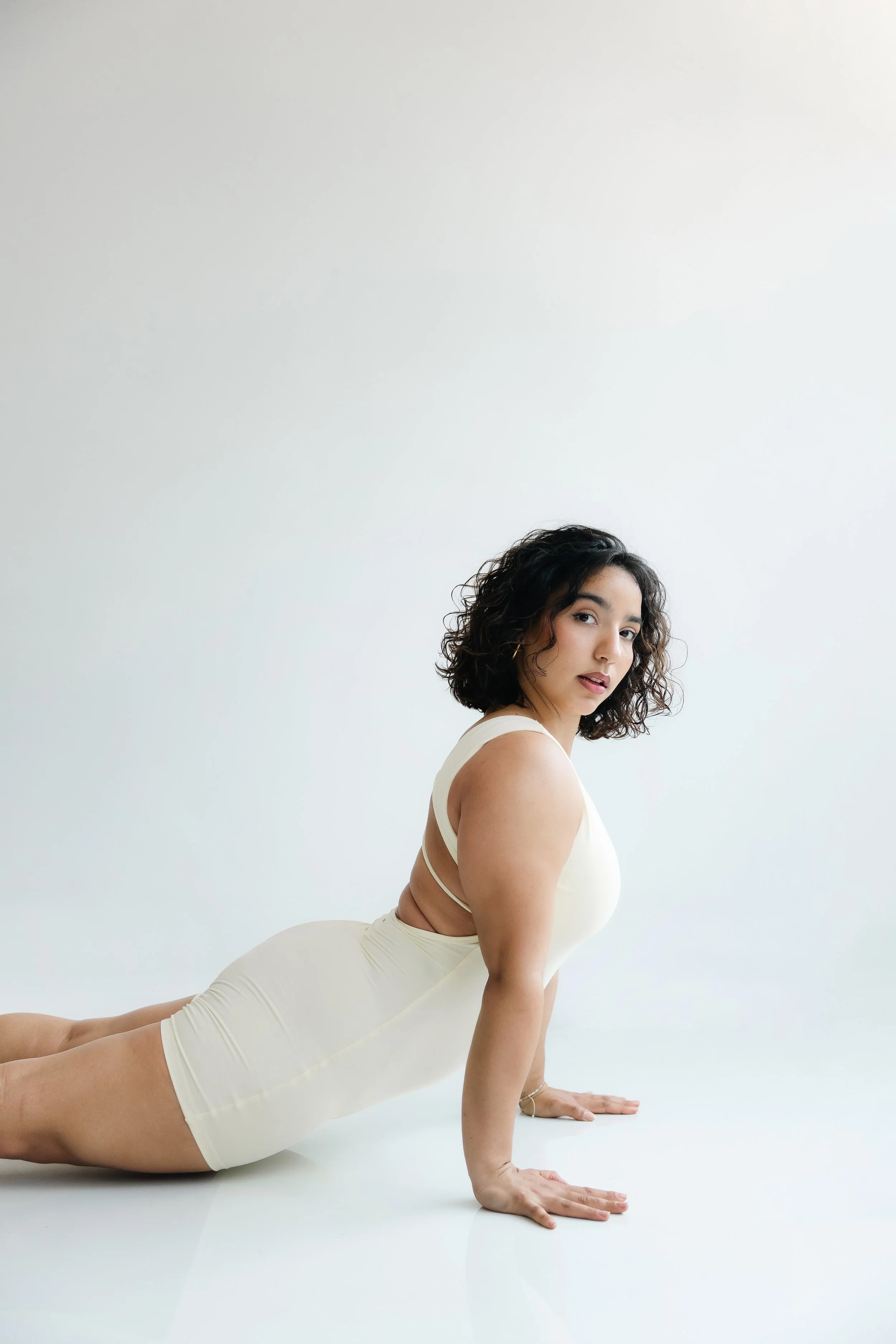 Woman in a beige dress doing a cobra yoga pose on a white floor against a plain white background.