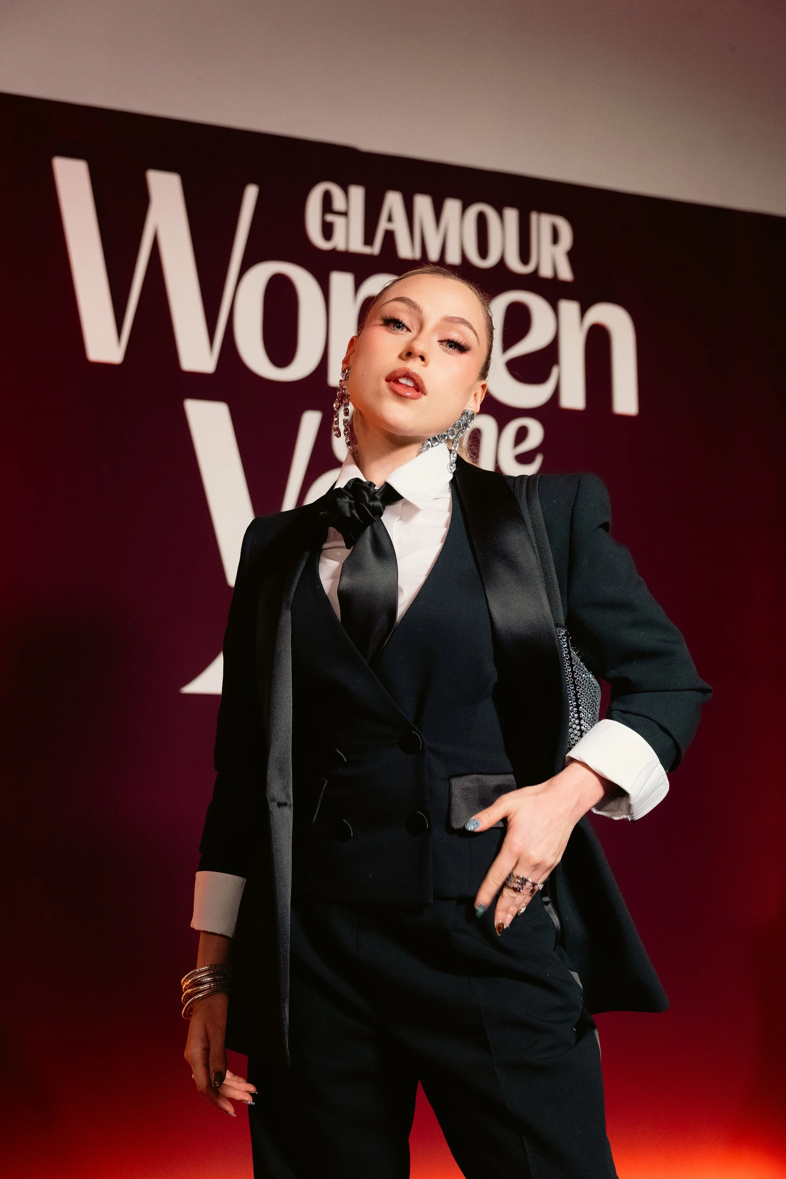 A woman in a black suit with a satin tie and white shirt standing in front of a Glamour Women of the Year sign.