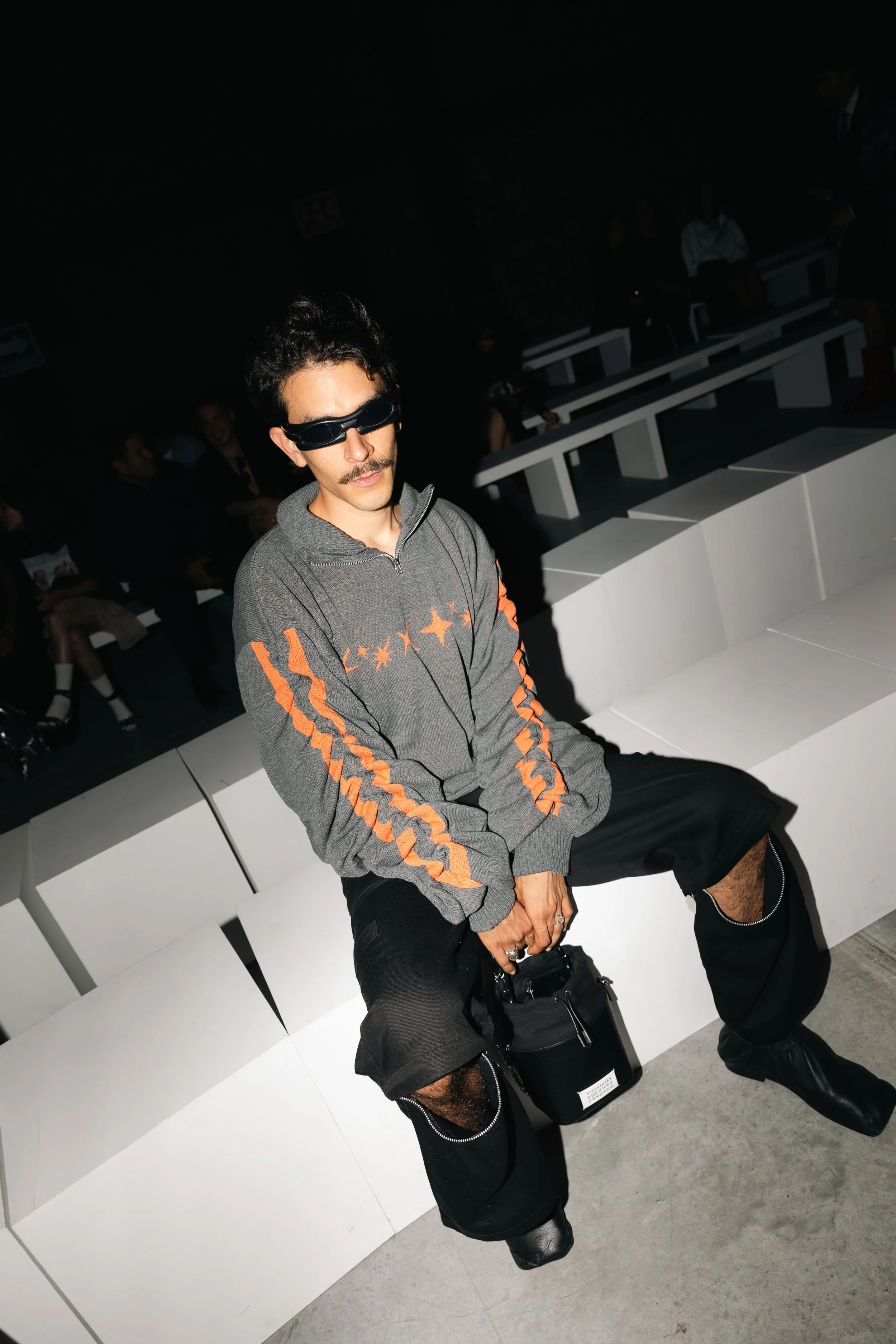 A man with dark hair and a mustache wearing black sunglasses, a grey zip-up jacket with orange stripes and star patterns, black pants with zippers on the knees, and black boots. He is sitting on a white bench in a dimly lit room with other people in 
