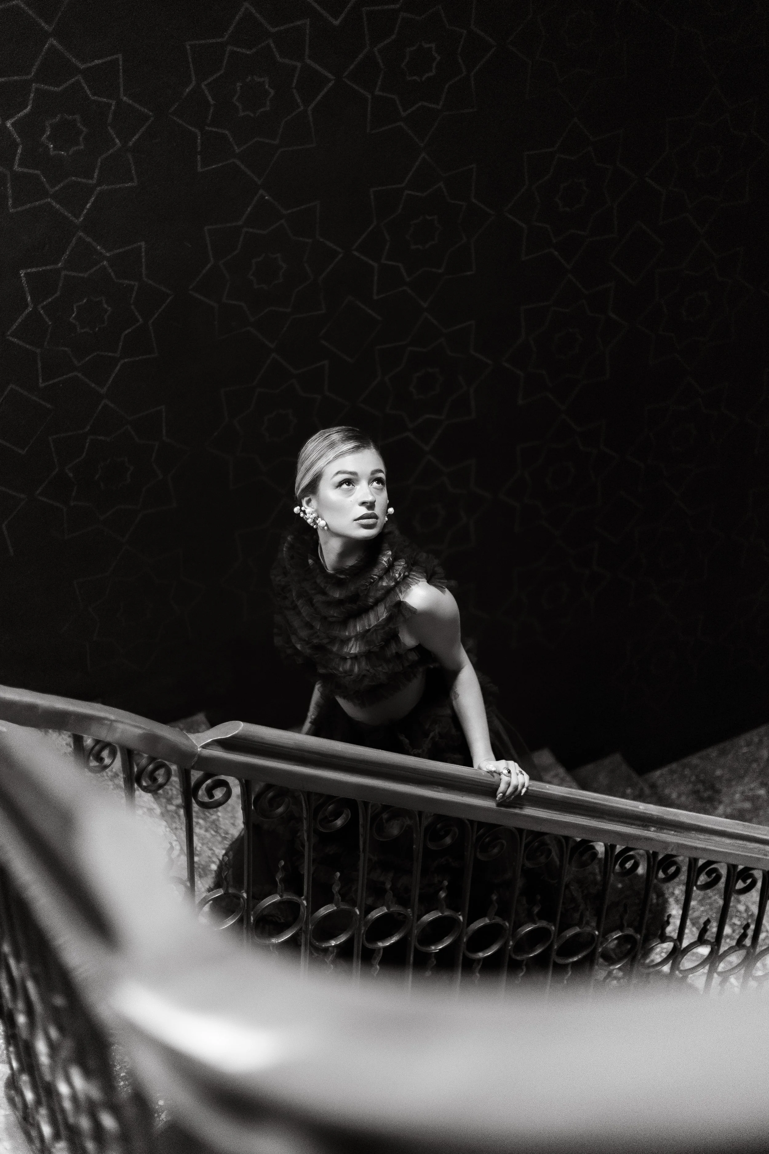 A woman with styled hair and wearing earrings, holding a staircase railing, looking up in a black-and-white photo with geometric wall patterns in the background.
