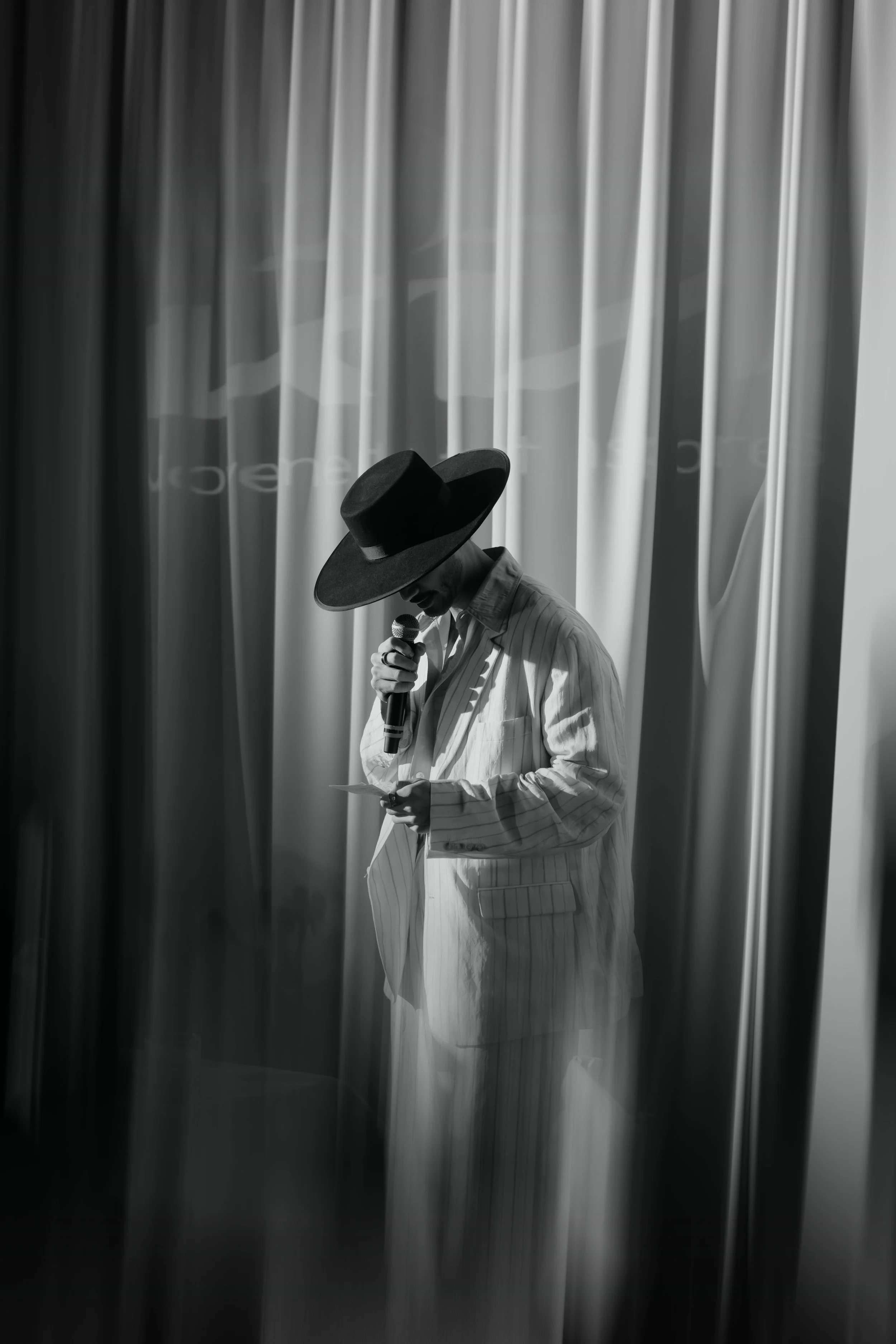 A person in a striped suit and large hat reading a paper and speaking into a microphone on stage with curtains in the background.