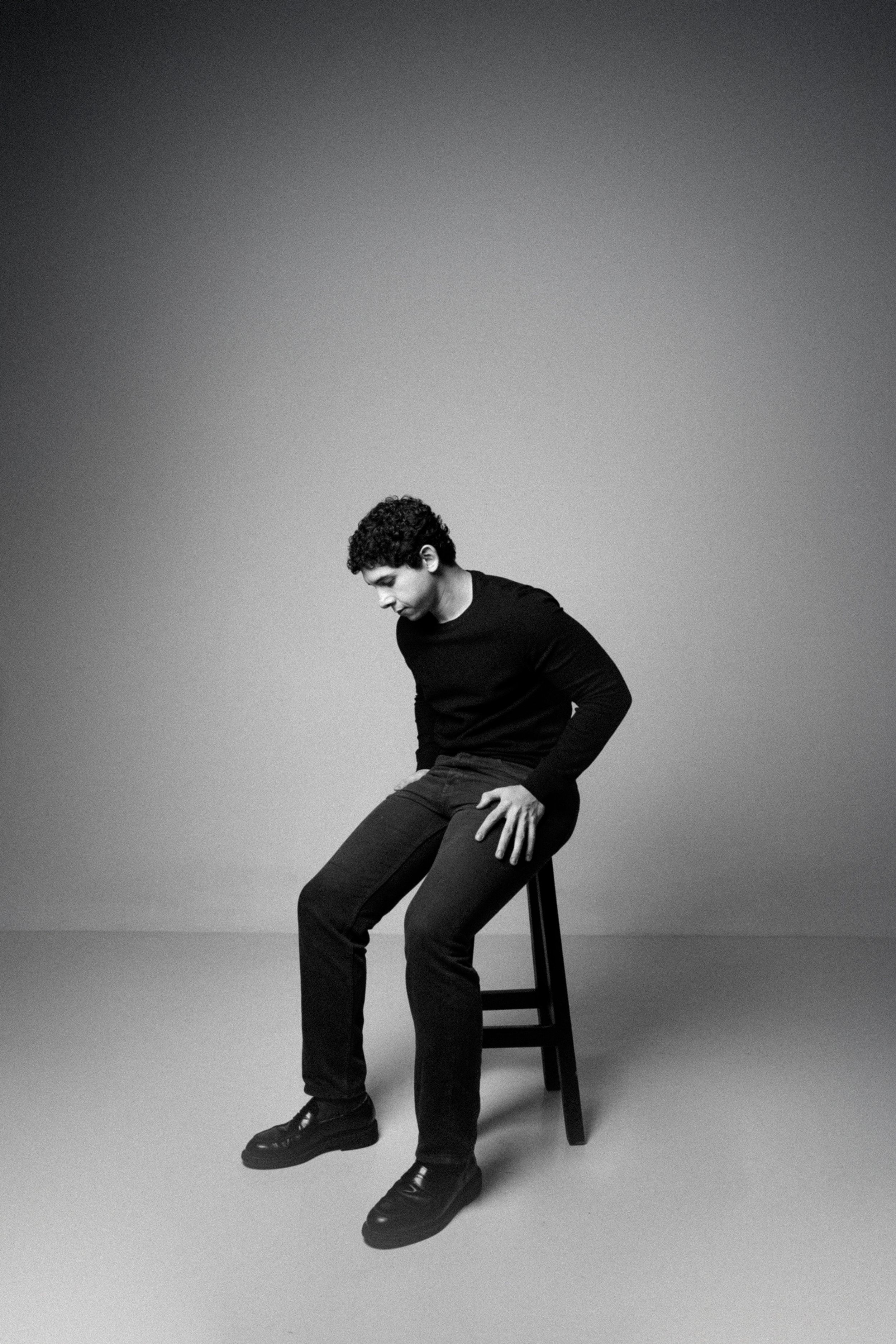 A young man with curly hair is sitting on a chair in a minimalist studio with a plain background. He is wearing a black long-sleeve shirt, dark pants, and black shoes, and is looking down with a contemplative expression.
