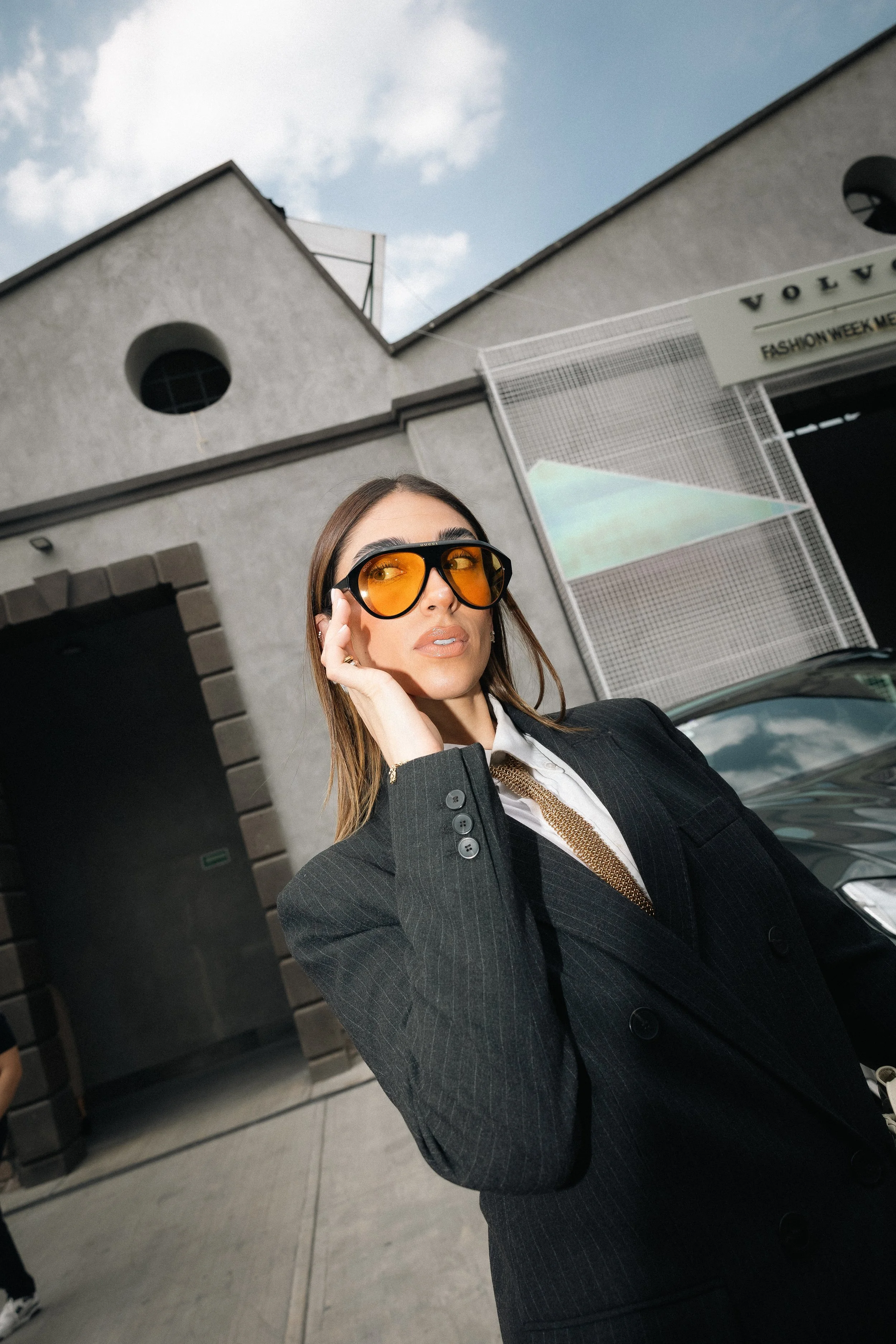 A woman in a black blazer and white shirt wearing oversized orange sunglasses stands outdoors near a building with a sign that says 'VOLVO' during the daytime, with cloudy sky above.