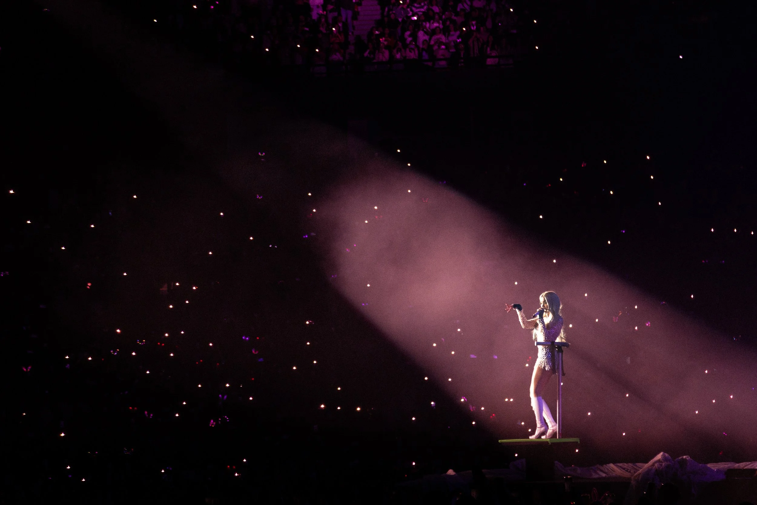 A performer on stage in a white outfit, illuminated by a spotlight, during a concert at night, with a dark background and many small pink and purple lights in the audience.