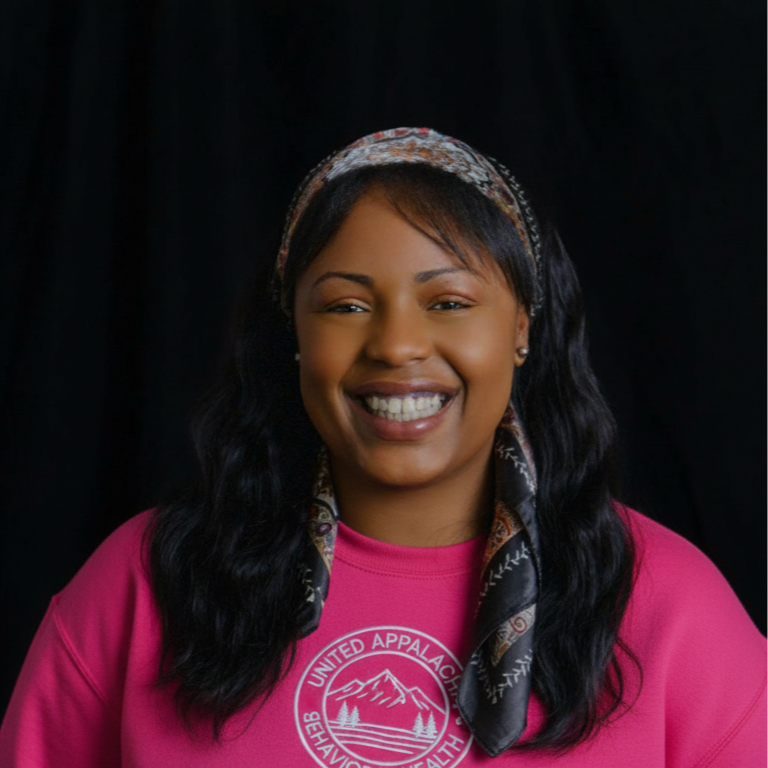 Smiling woman with dark hair, wearing a pink shirt and a patterned headband, against a black background.