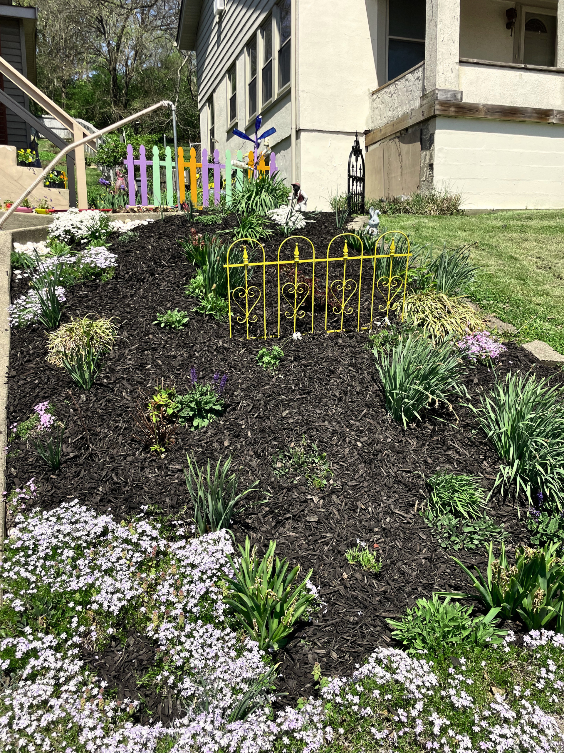 Colorful garden bed with flowers, small decorative fences, and garden ornaments outside a house.