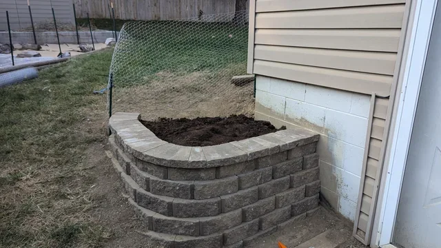 Newly constructed raised garden bed made of gray bricks, located next to the house's exterior wall, with dark soil inside.