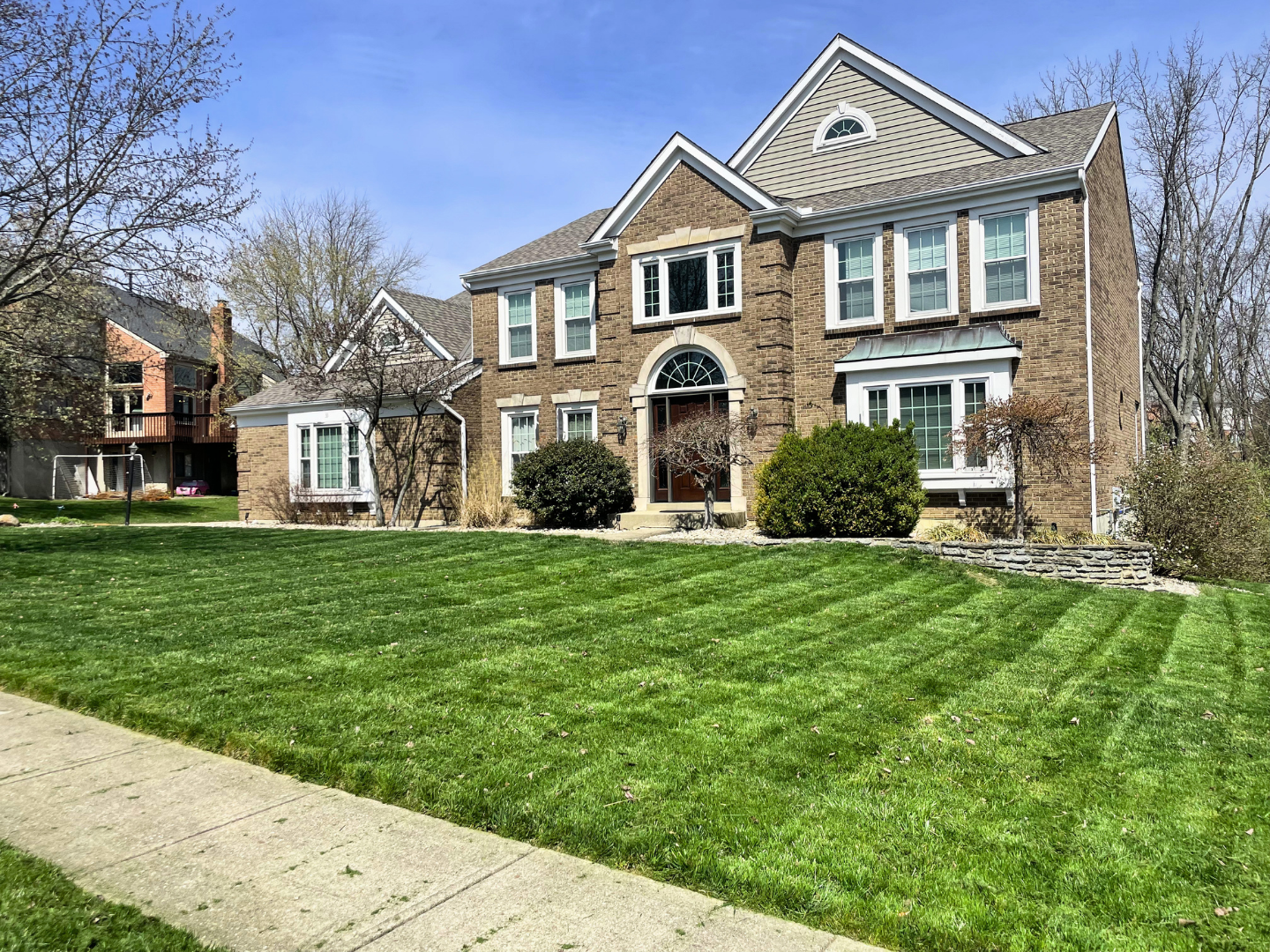 A large brick house with a well-maintained green lawn and mature trees, under a partly cloudy blue sky.