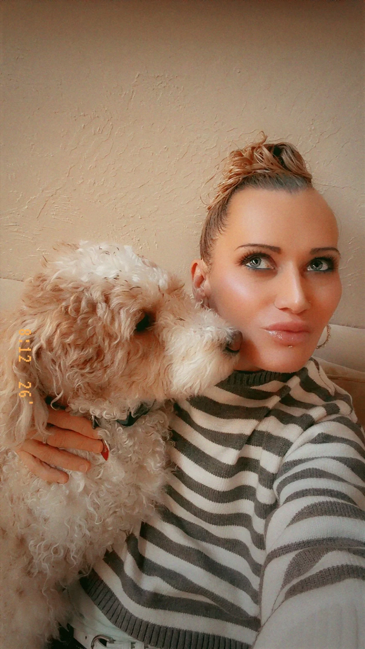 A woman with light skin, blue eyes, and styled hair in a bun holding and possibly kissing a curly-haired dog with light-colored fur.