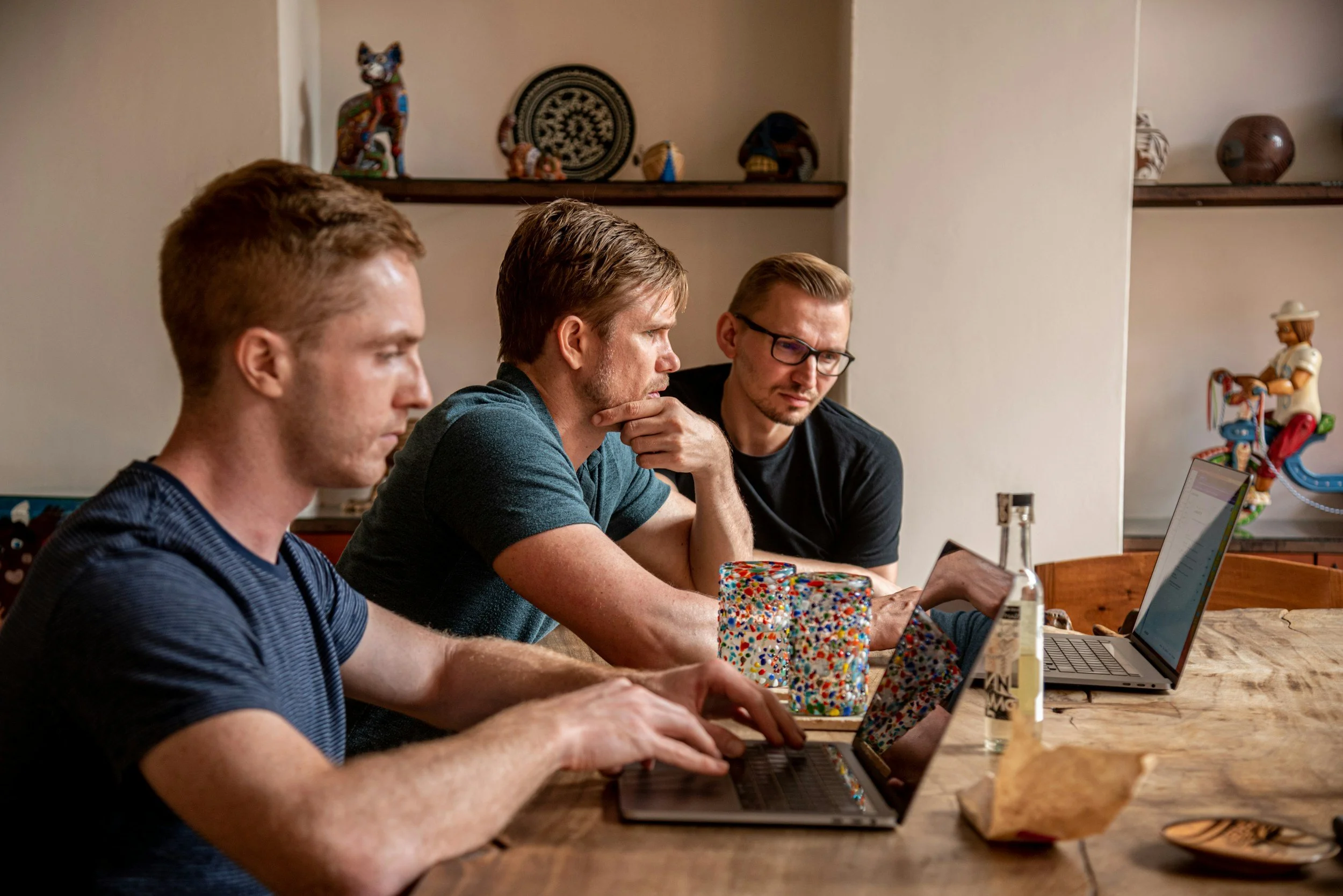 Three men sitting at a wooden table working on laptops, with colorful decorations and small sculptures in the background.