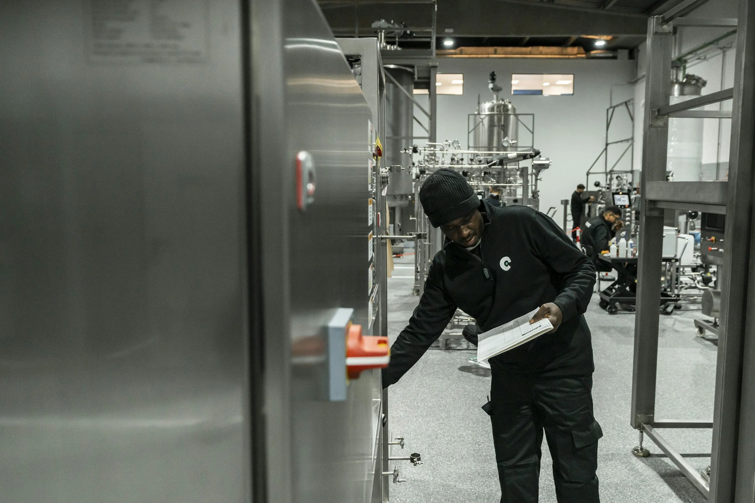 A man in a black beanie and jacket holding a notebook examines industrial equipment in a modern, well-lit manufacturing or laboratory facility with machinery and other workers in the background.
