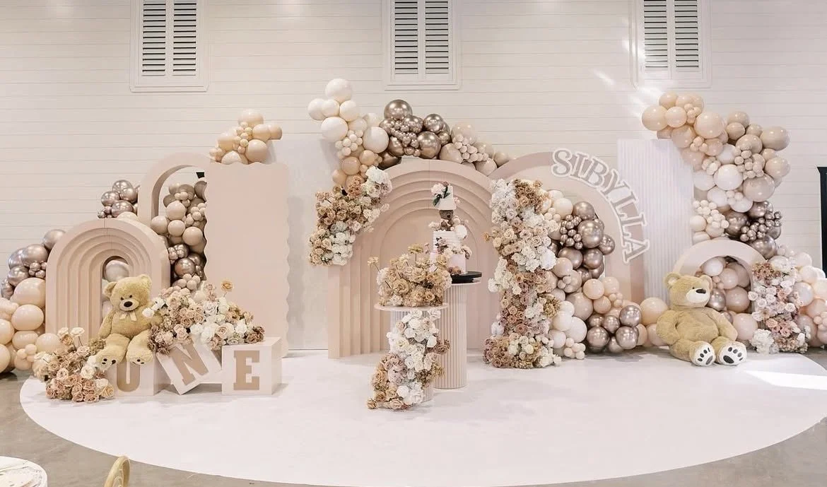 Decorative backdrop with pink, white, and silver balloons, flowers, and teddy bears, likely for a first birthday celebration.