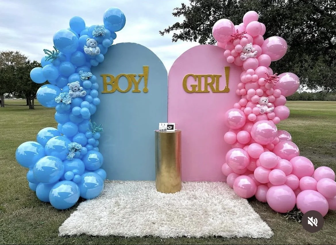 Decorative display with blue and pink balloon arrangements labeled Boy and Girl, with teddy bears and flowers, set outdoors on a grassy field.